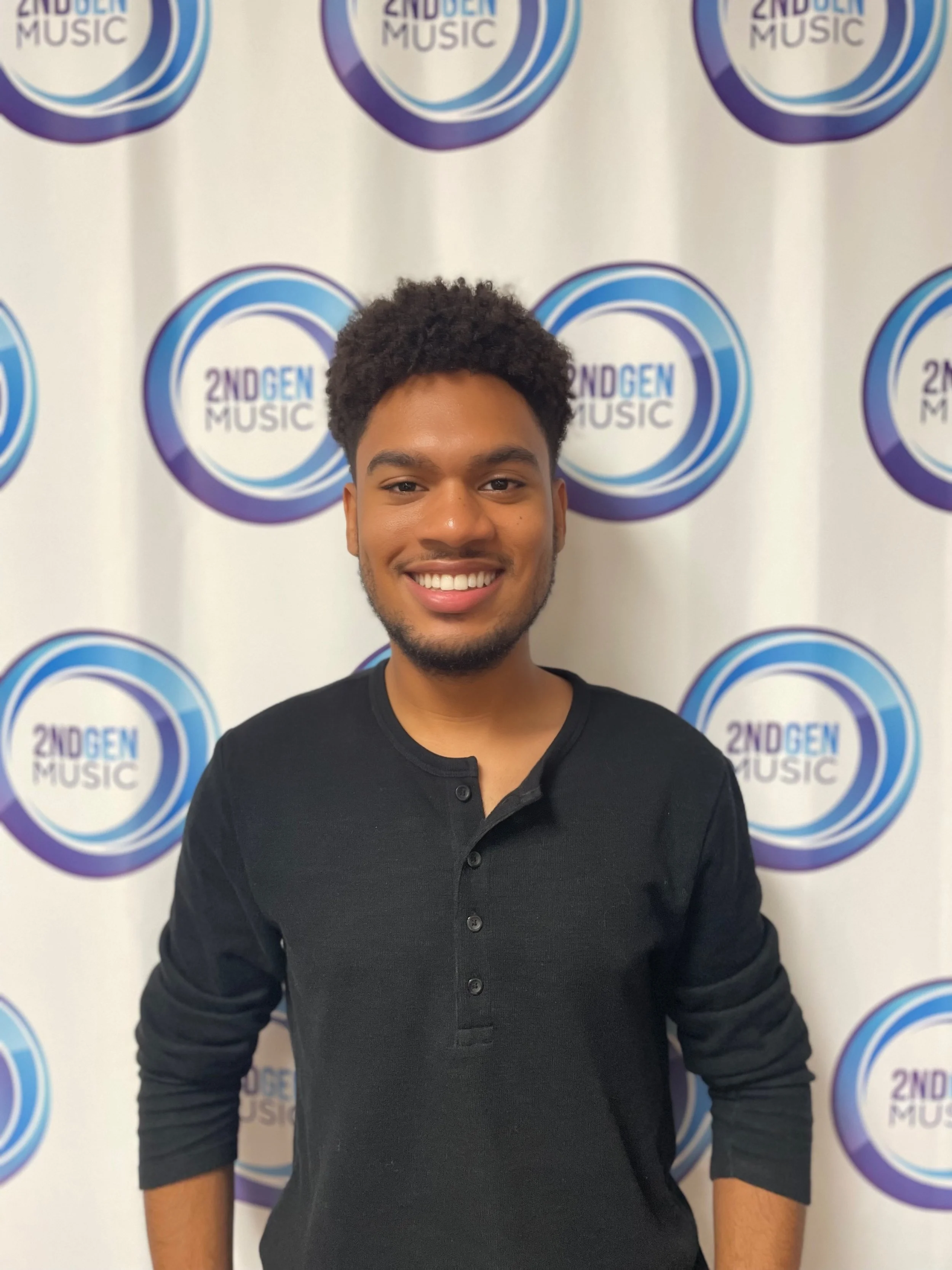 A young man with curly hair and a beard smiling, standing in front of a backdrop with a logo that reads '2ND GEN MUSIC' repeatedly.