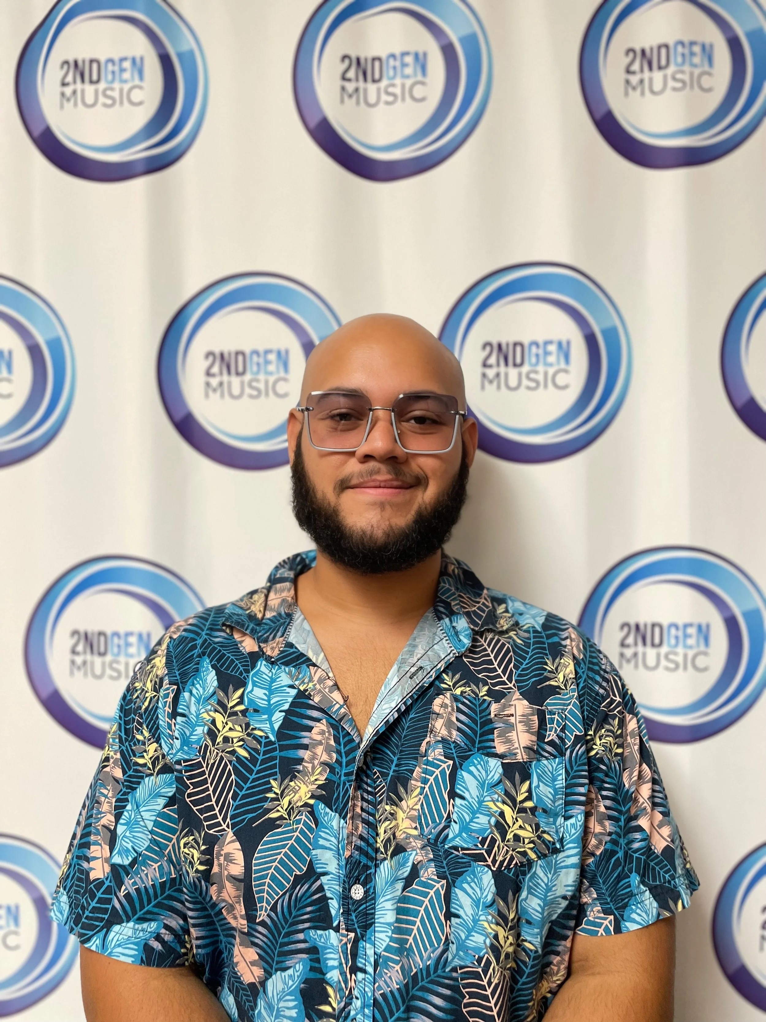 A man with a beard and glasses wearing a colorful tropical shirt, standing in front of a backdrop with multiple '2ND GEN MUSIC' logos.