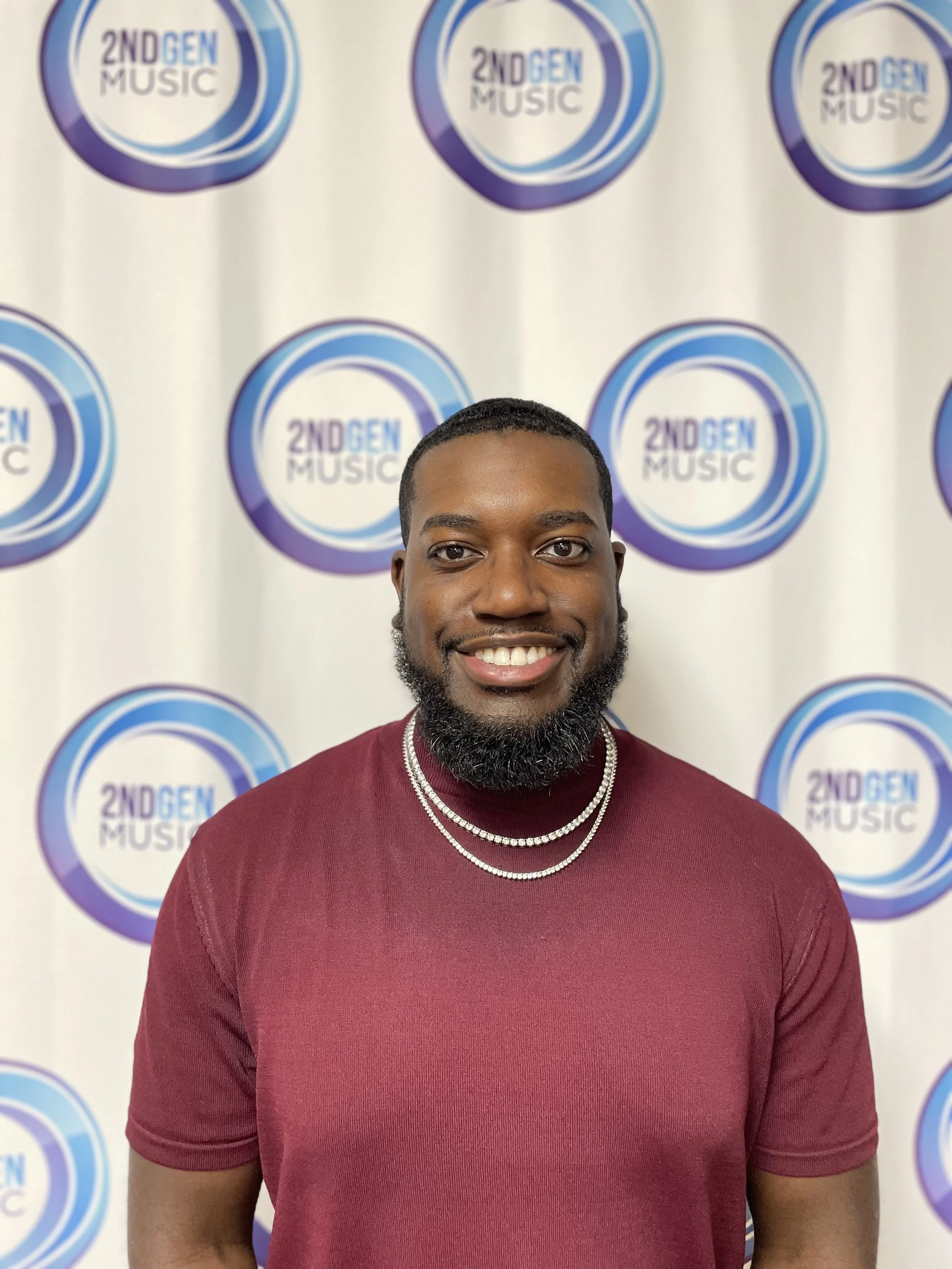 A smiling man with a beard, wearing a maroon shirt and layered silver necklaces, standing in front of a backdrop with multiple logos that read '2ND GEN MUSIC' in a circular design with blue and purple tones.