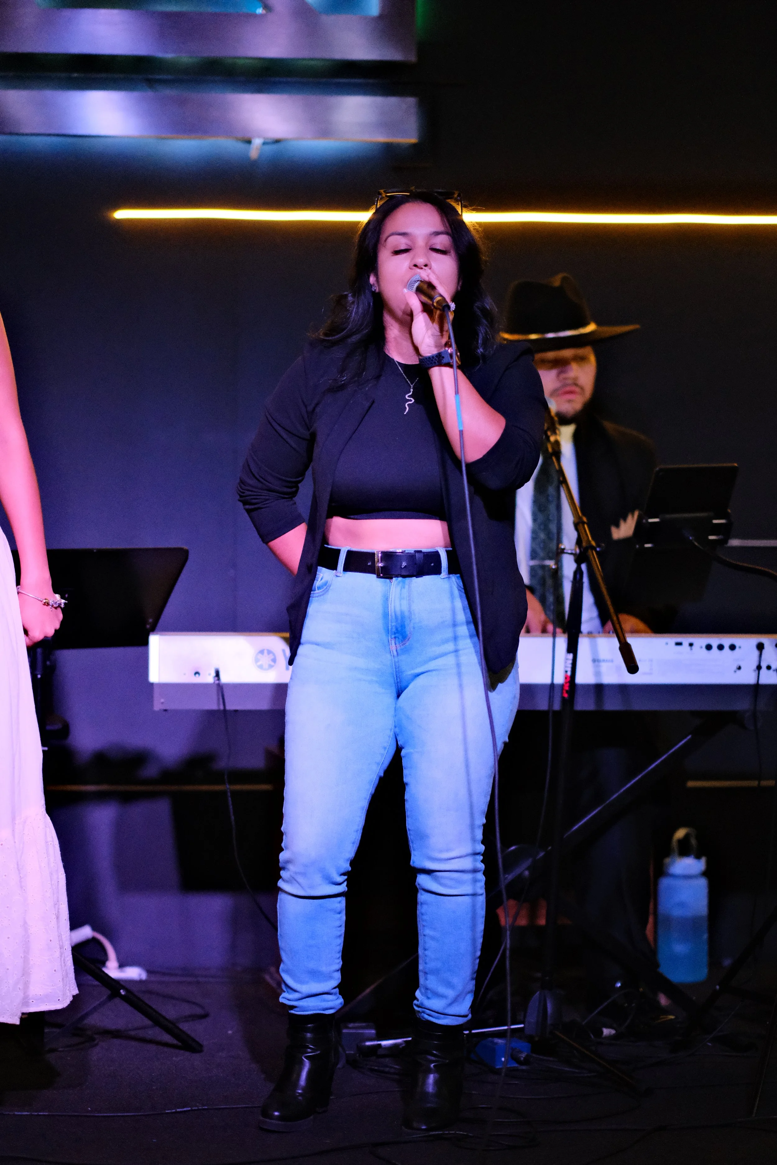 Woman wearing jeans and tank top holding a microphone singing with a man in a hat playing the piano.