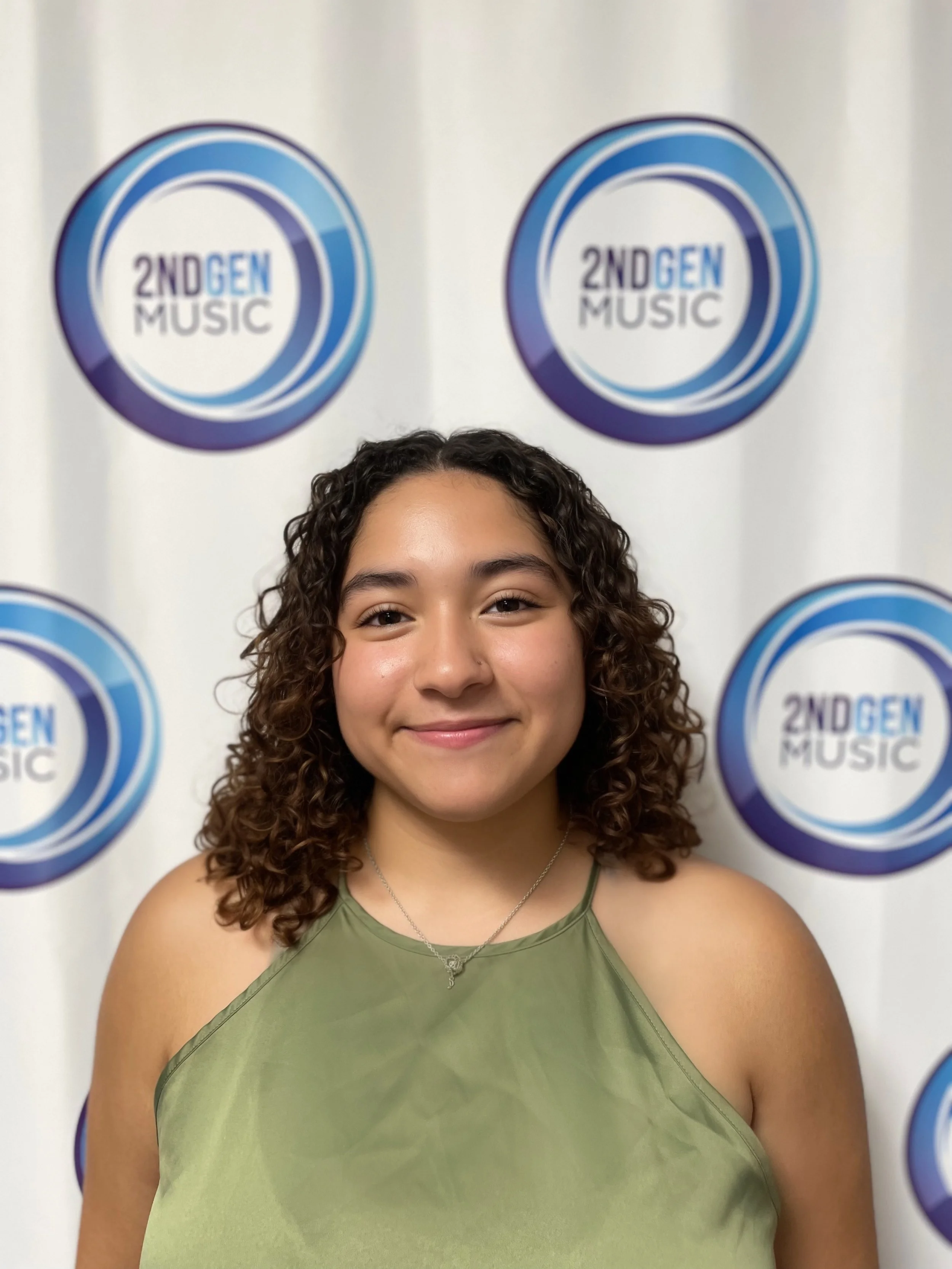 A young woman with curly hair, smiling, wearing a green sleeveless top, standing in front of a white backdrop with blue circular logos that say "2ND GEN MUSIC."