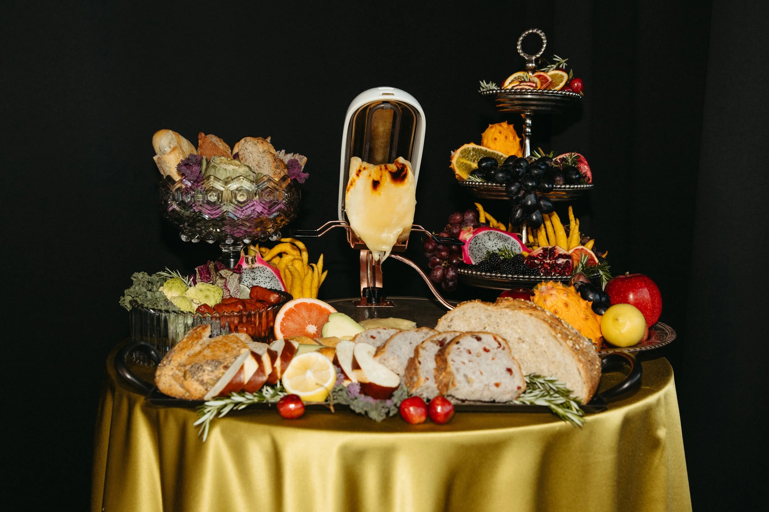 Table setting with cheese raclette melting under the heat of a raclette melter, a variety of breads, and assorted fruits accompany cheese.