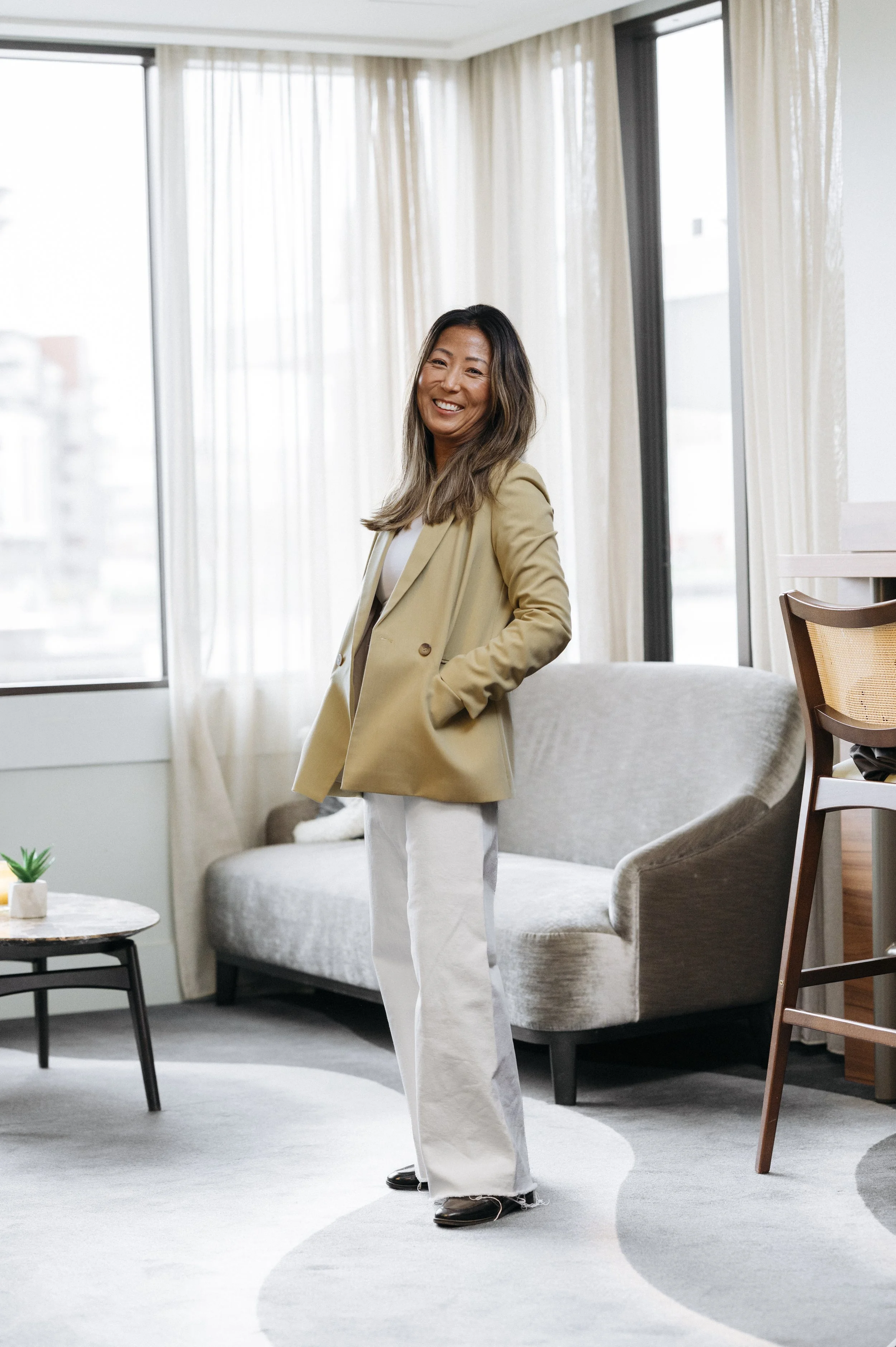 Therese Jung står smilende foran store vinduer, iført beige blazer og lyse bukser, med hånden i lommen.