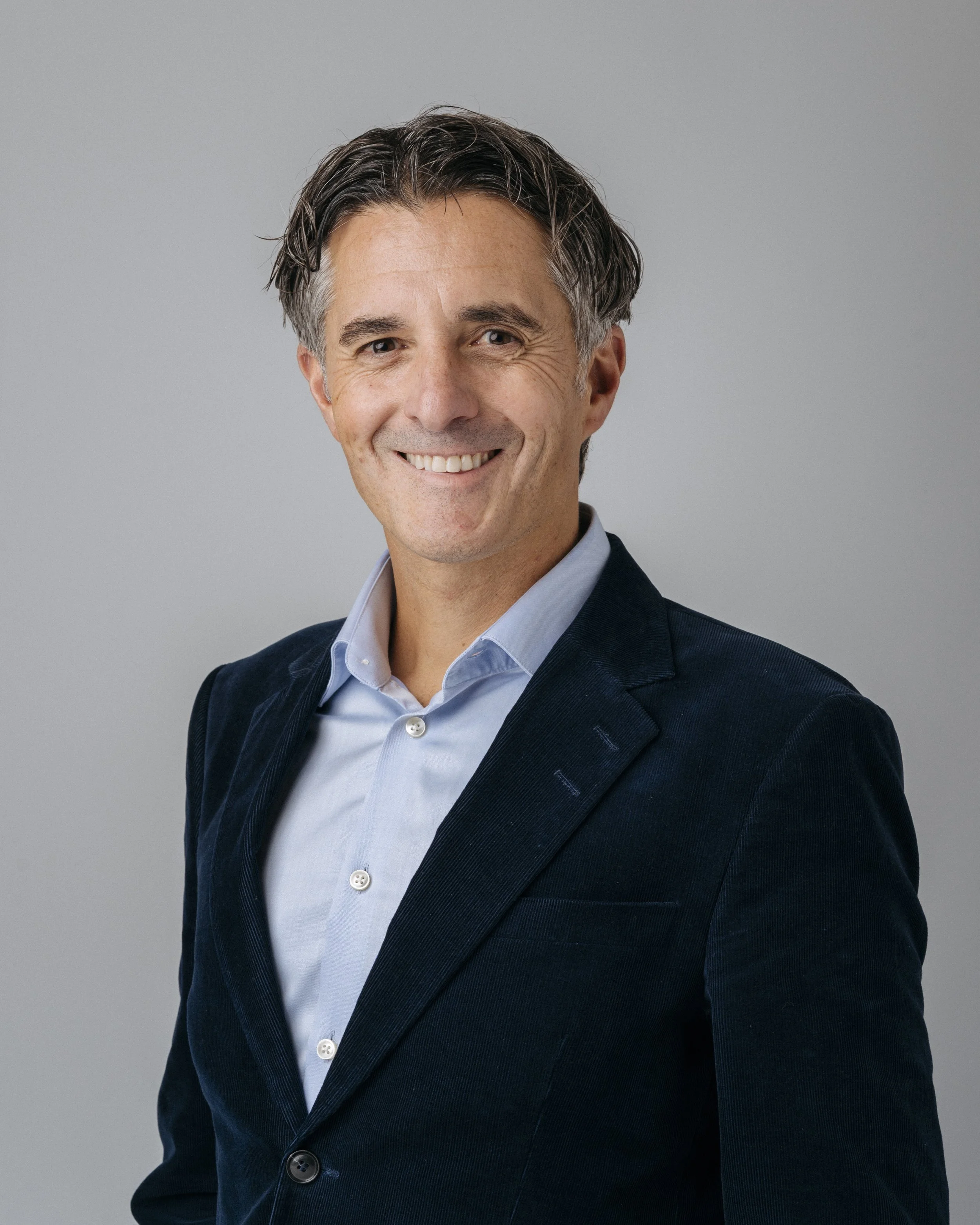 Et porträtt av en mann i dress, smilende mot kamera mot en grå bakgrunn.