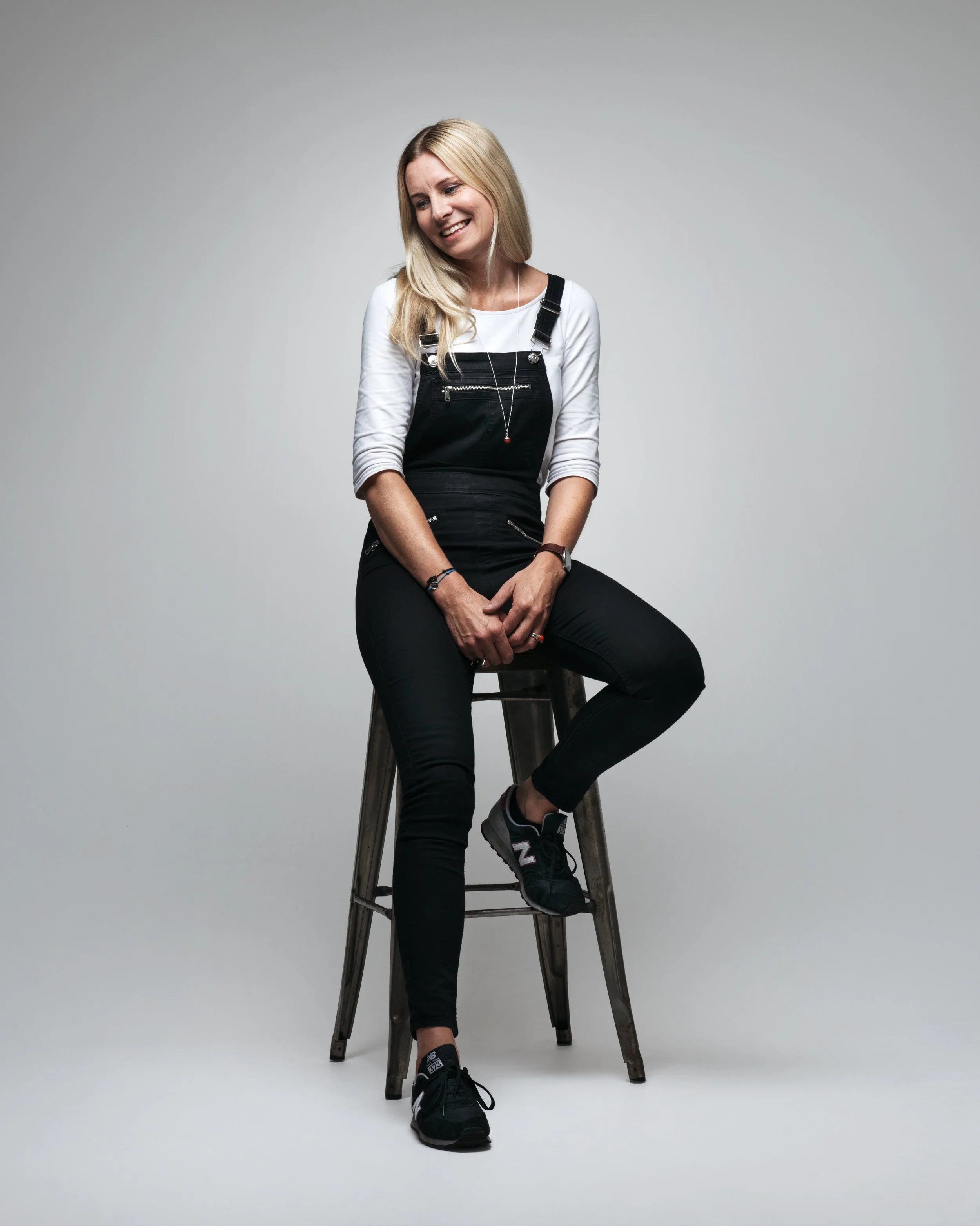 En smilende kvinne med langt blondt hår, iført svart buksedress over en hvit langermet skjorte, sitter på en skjenk mot en lys grå bakgrunn.