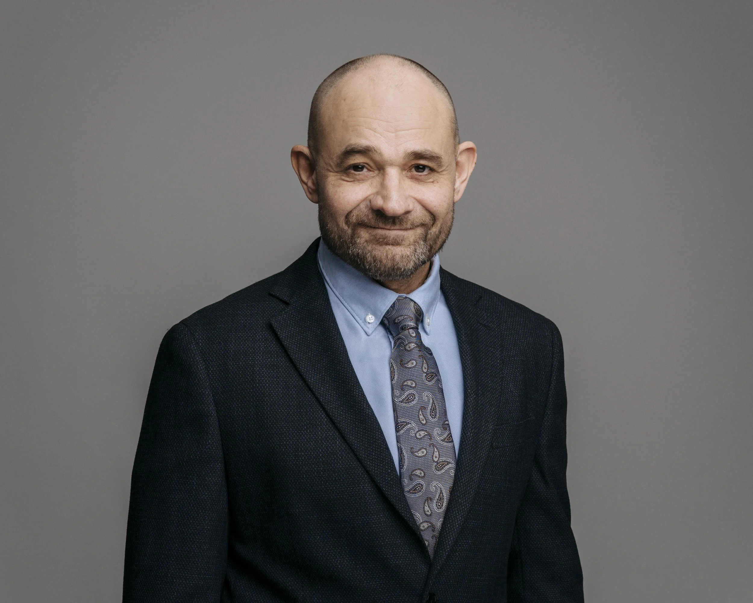 En mann i mørk dress og blå skjorte med slips, smilende mot kamera, stående mot en grå bakgrunn.