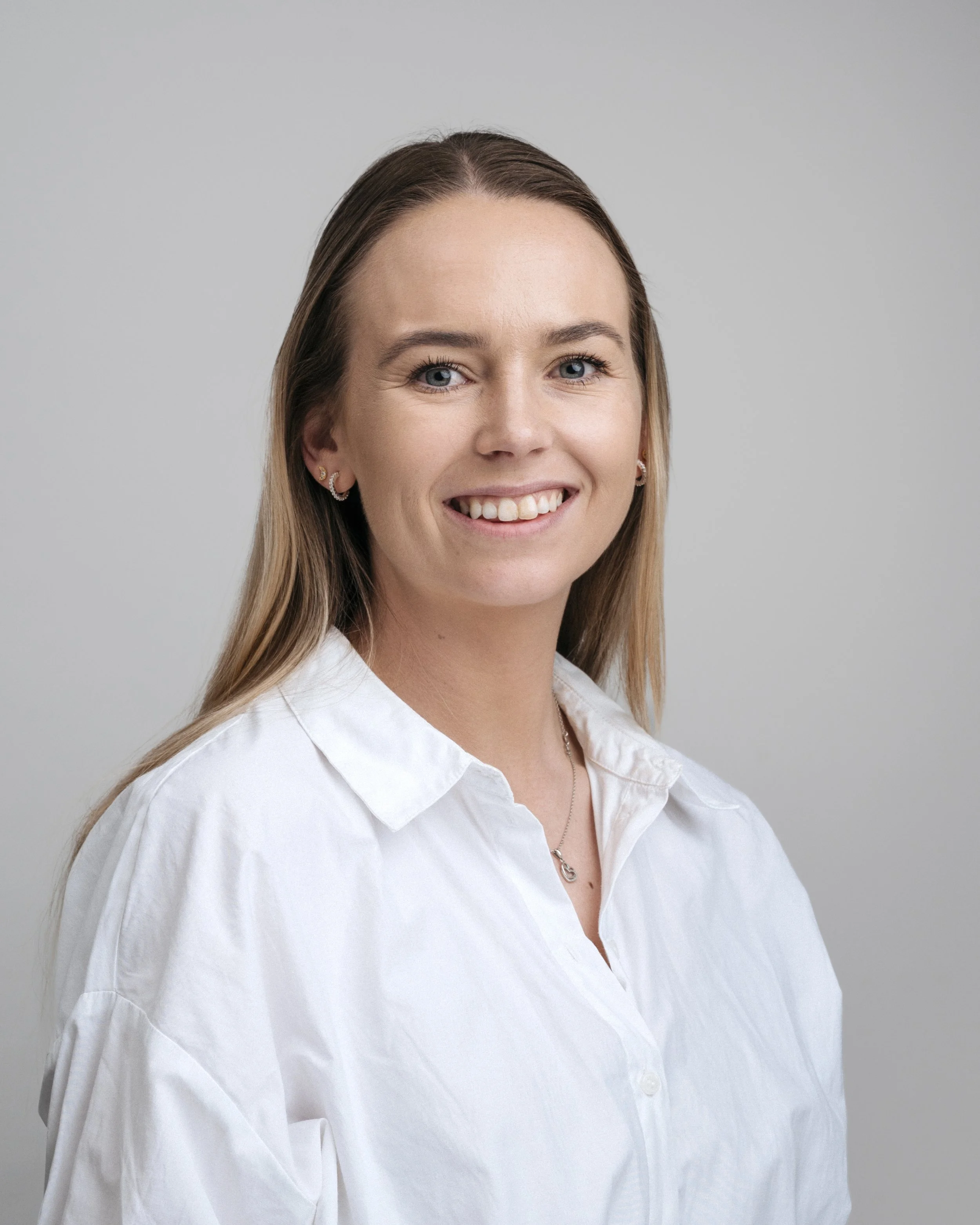 En kvinne med langt blondt hår, smilende, iført hvit skjorte og smykker, mot en nøytral grå bakgrunn.