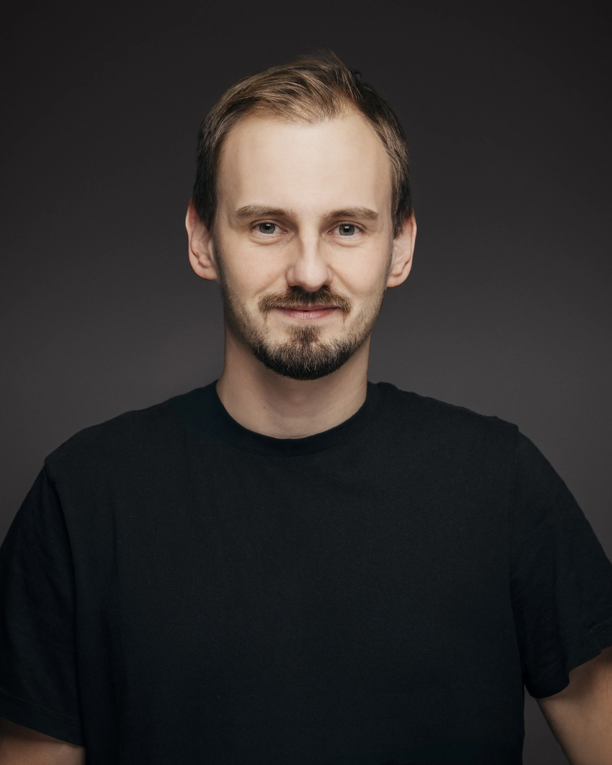 Portrett av en ung mann med lysebrunt hår, skjegg, iført svart T-skjorte, mot en mørk bakgrunn.