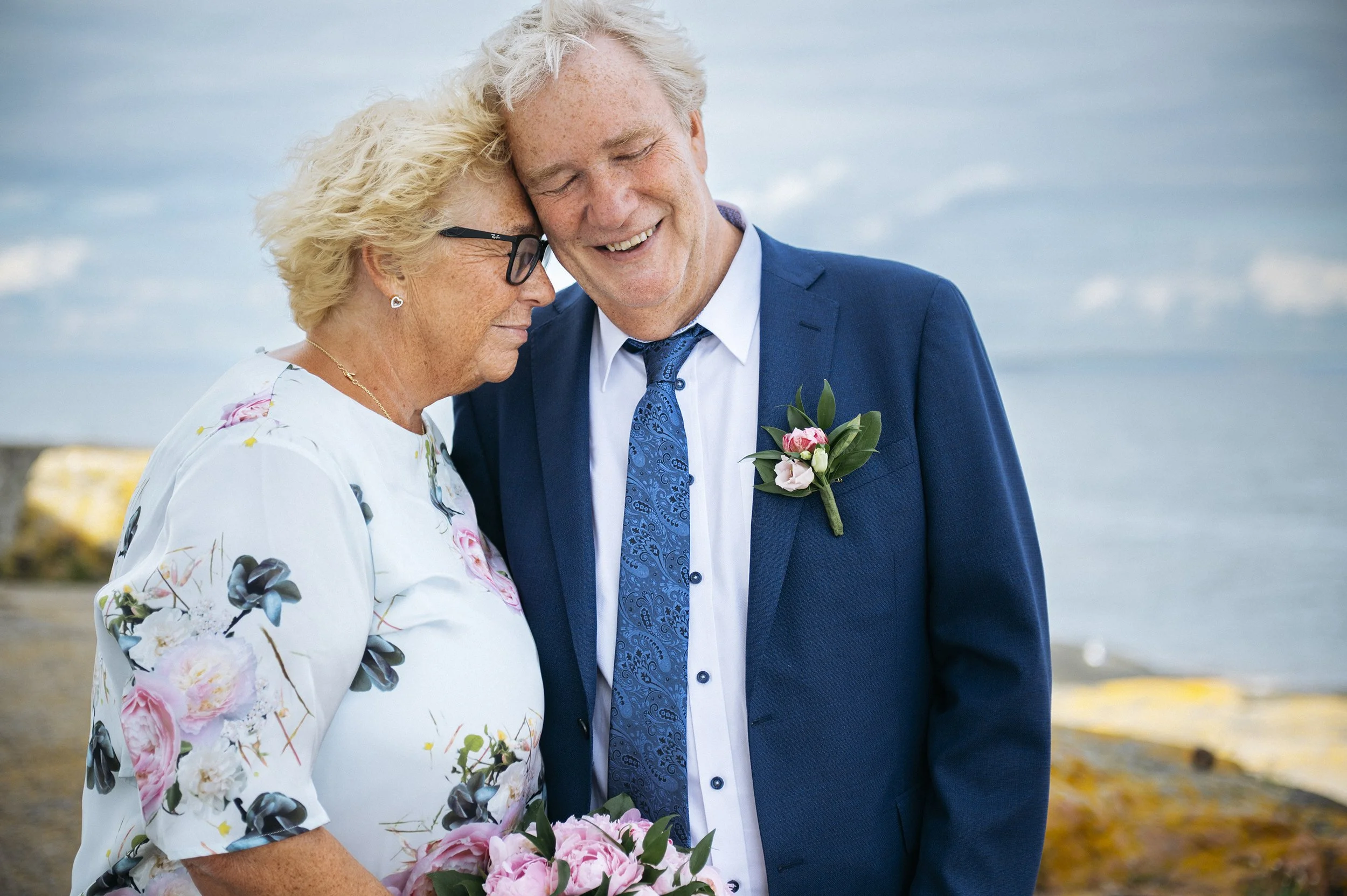 Ektepar står tett sammen og smiler ved sjøen, et varmt og naturlig bryllupsportrett i et norsk kystlandskap på Fulehuk.