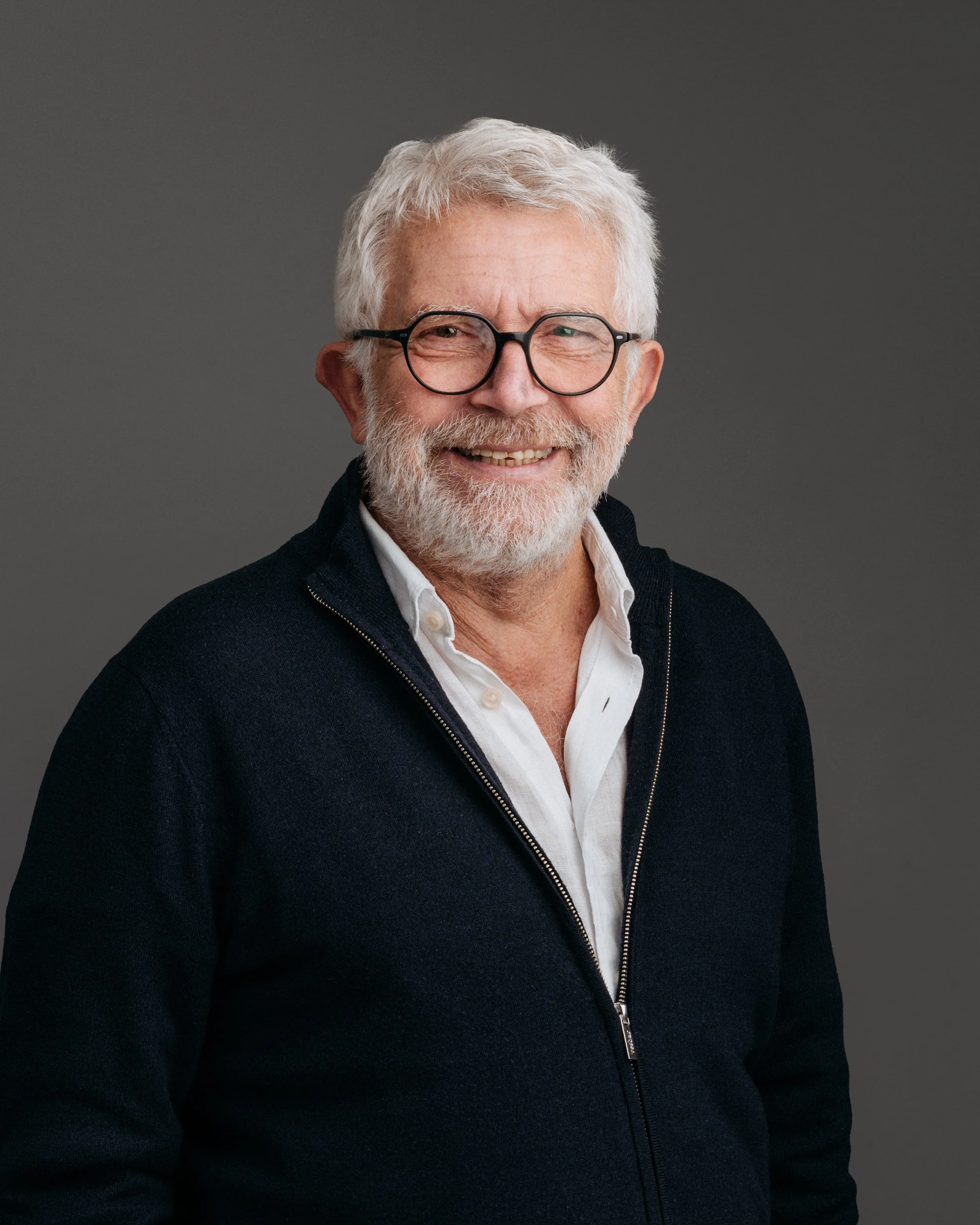 Portrett av en smilende eldre mann med grått hår, skjegg og briller, kledd i en hvit skjorte og mørk genser, mot en grå bakgrunn.