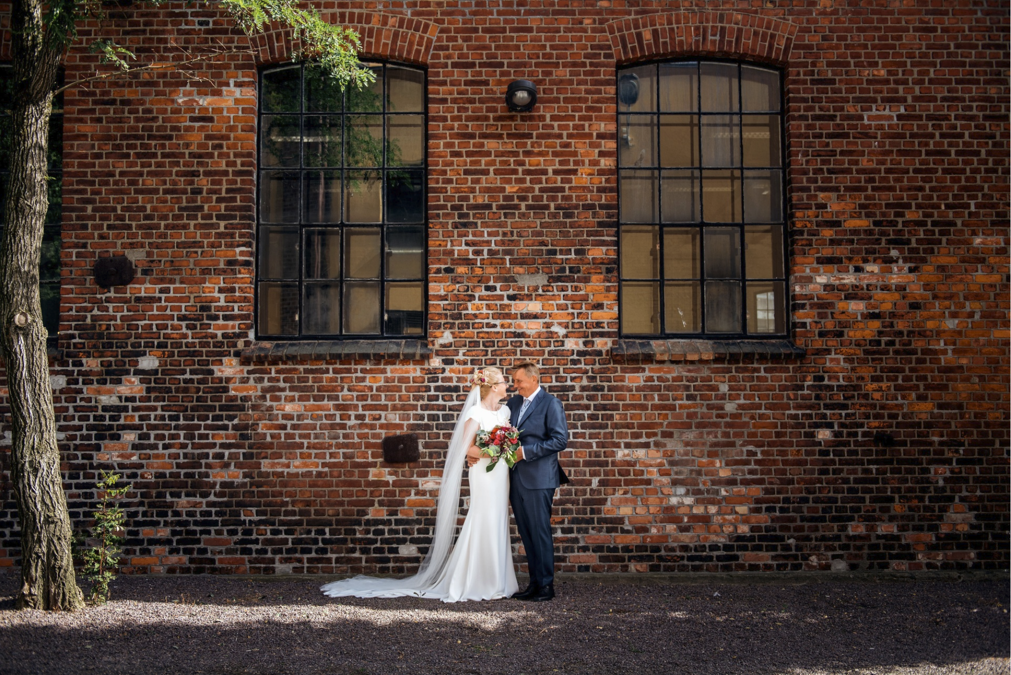En brud og en brudgom står sammen foran en rød murvegg. Bruden har på seg en hvit kjole og holder en blomsterbukett, og brudgommen har på seg en blå dress.