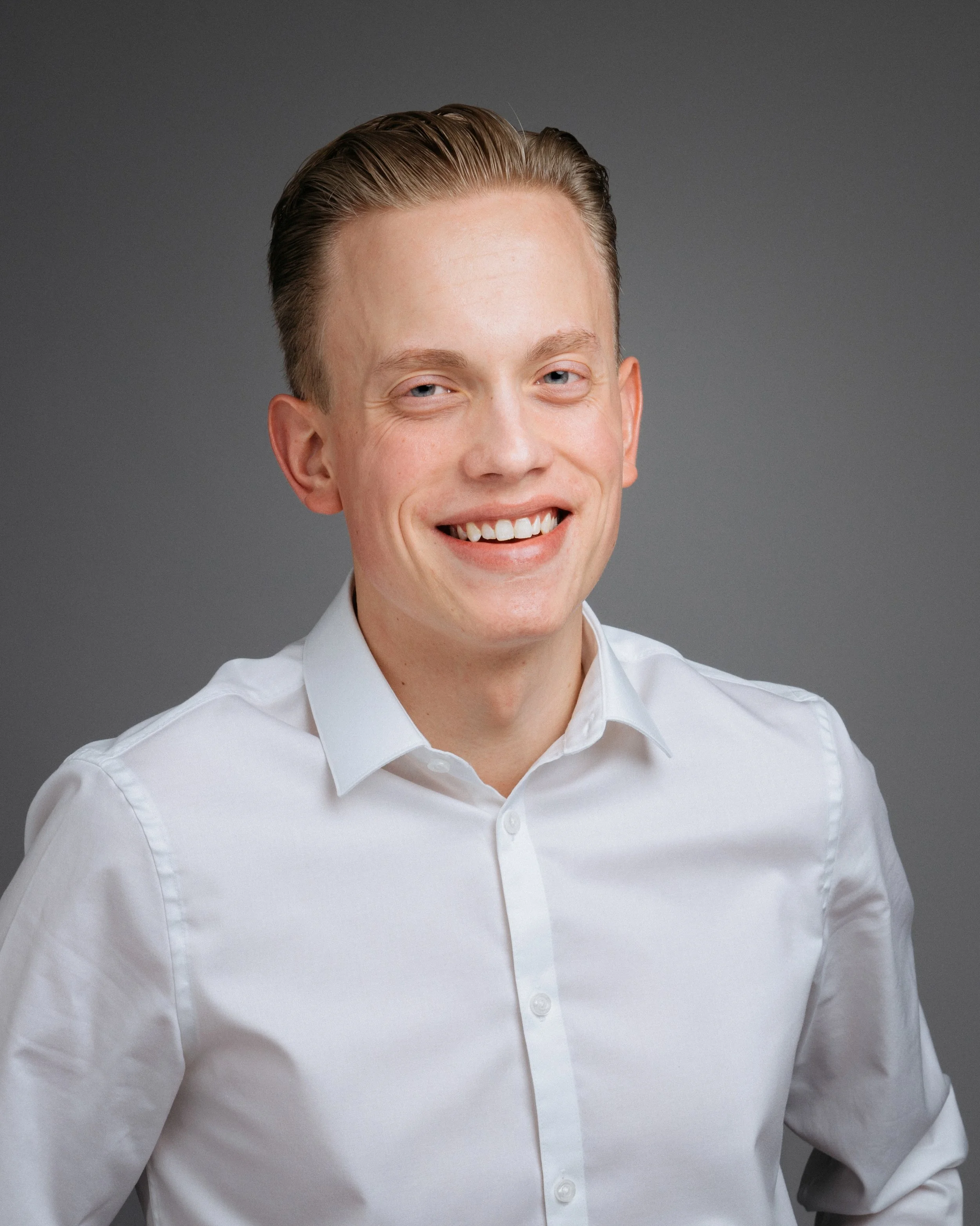 Ung mann med lys hud, blond hår, iført hvit skjorte, smilende mot kamera mot en grå bakgrunn.