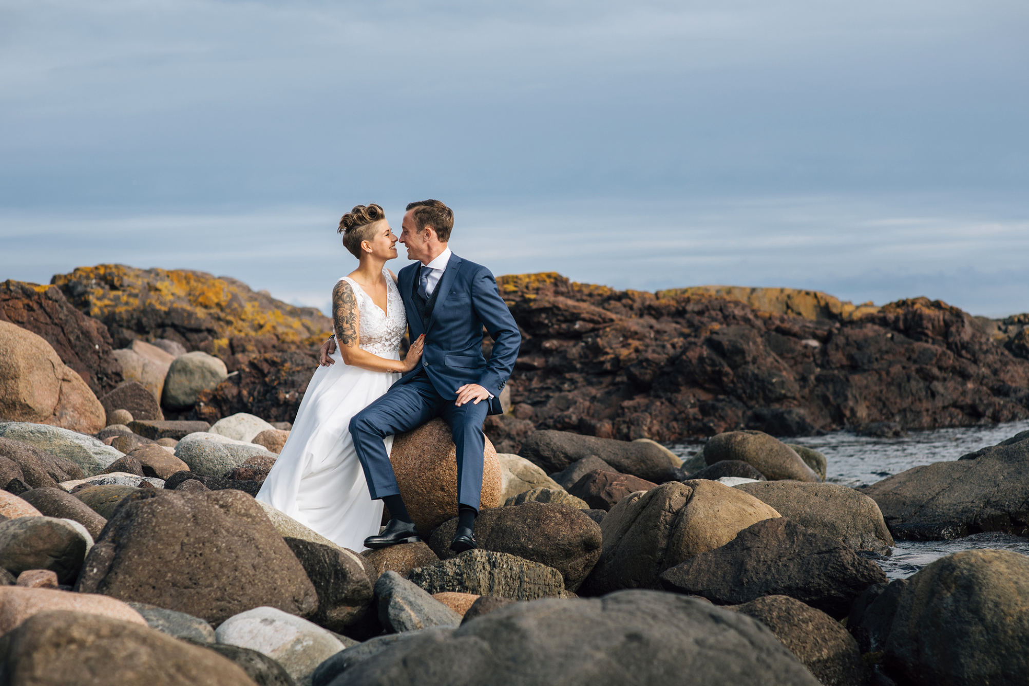 Brudepar sitter tett sammen på svaberg ved havet, et romantisk bryllupsportrett i et rått og nordisk kystlandskap på Karlsvika i Tønsberg.
