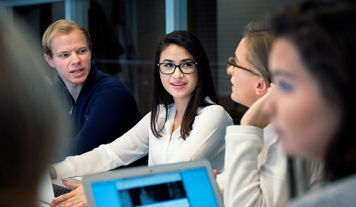Miljøfoto av studenter som sitter samlet rundt et bord i en samtale- eller arbeidssituasjon.