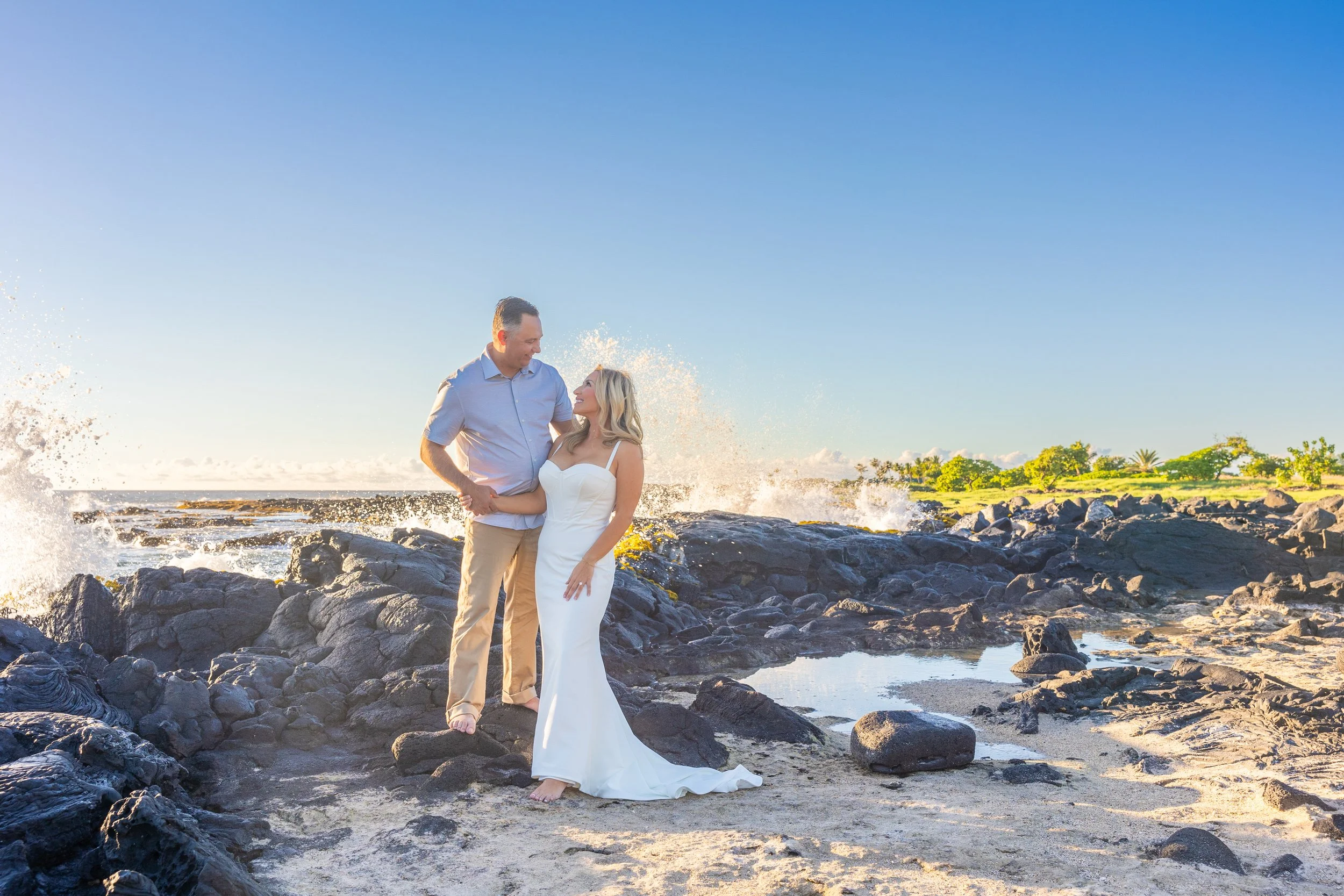 A couple in wedding attire stands on black rocks by the ocean with waves crashing behind them, under a clear blue sky, during sunset or sunrise.