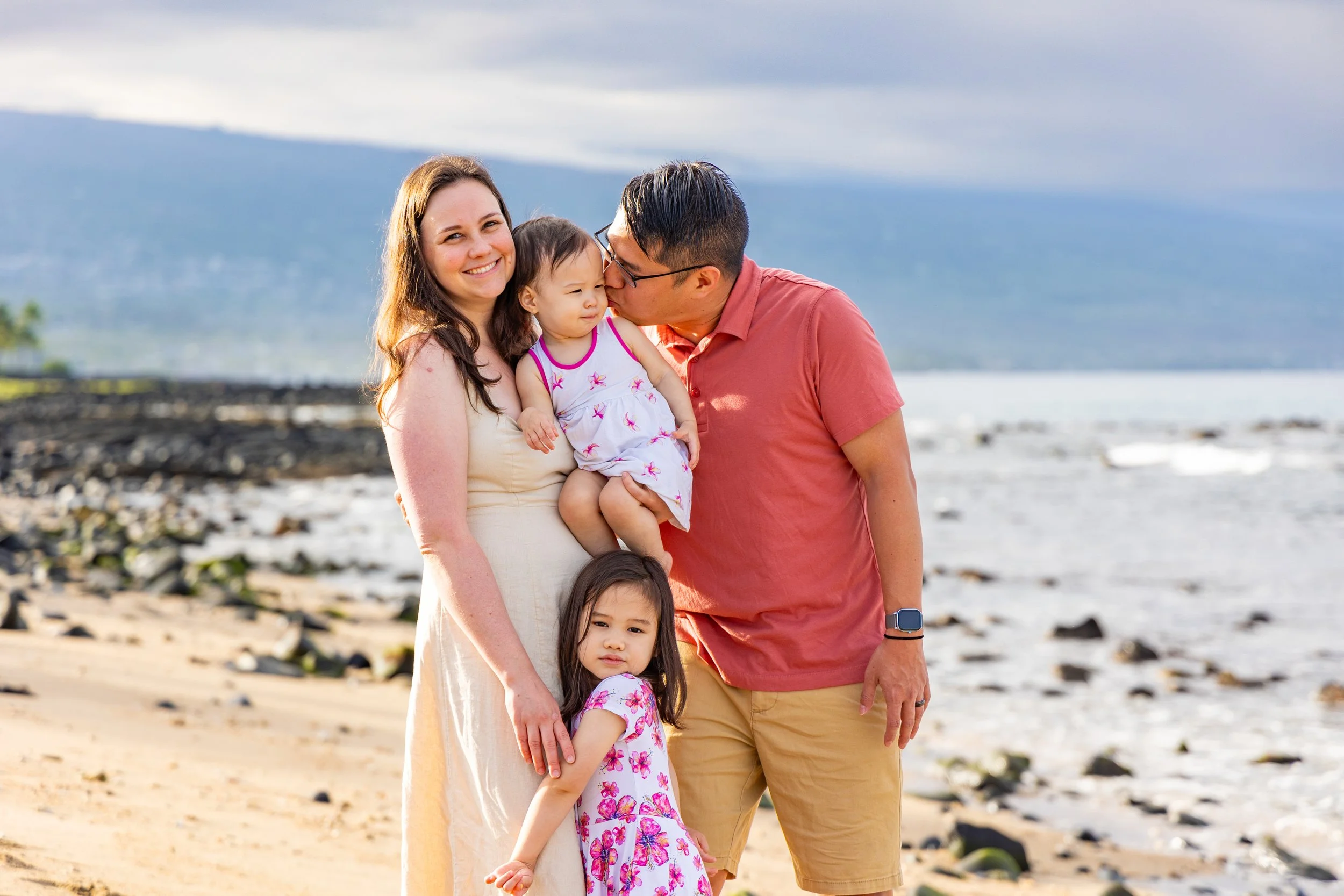 A family of four on a beach, with the mother holding a young girl and the father kissing a toddler girl on the cheek, the older girl standing in front of the mother, and the family smiling.