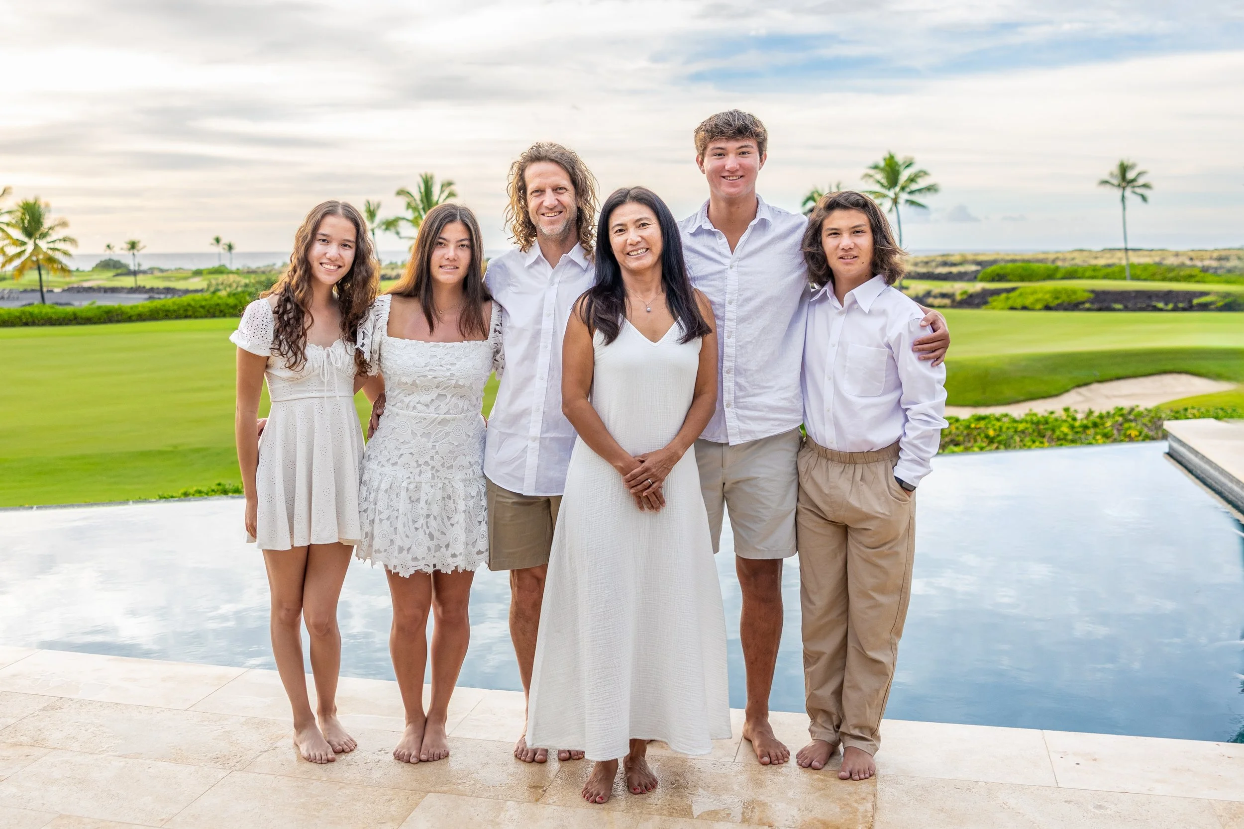 A family of seven dressed in white, standing together on a patio near a swimming pool with a golf course and palm trees in the background.