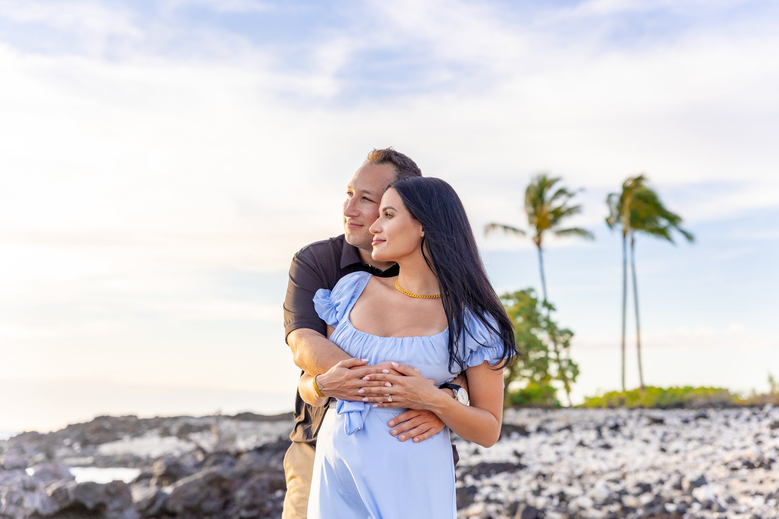 A couple stands on a rocky shoreline embracing, with wind-blown palm trees and a cloudy sky in the background.
