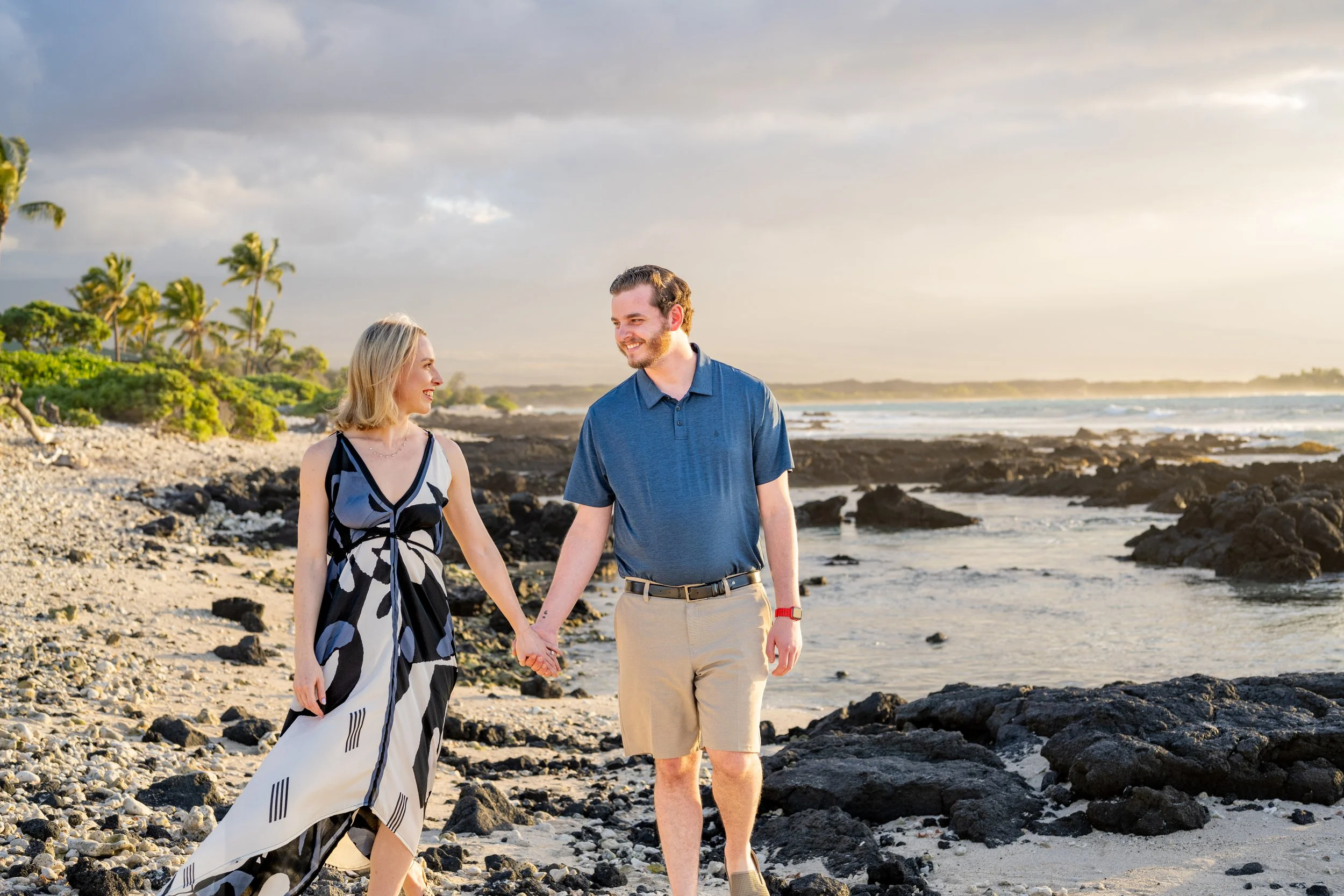 A happy couple walking hand in hand on a rocky beach during sunset, with palm trees and ocean in the background.