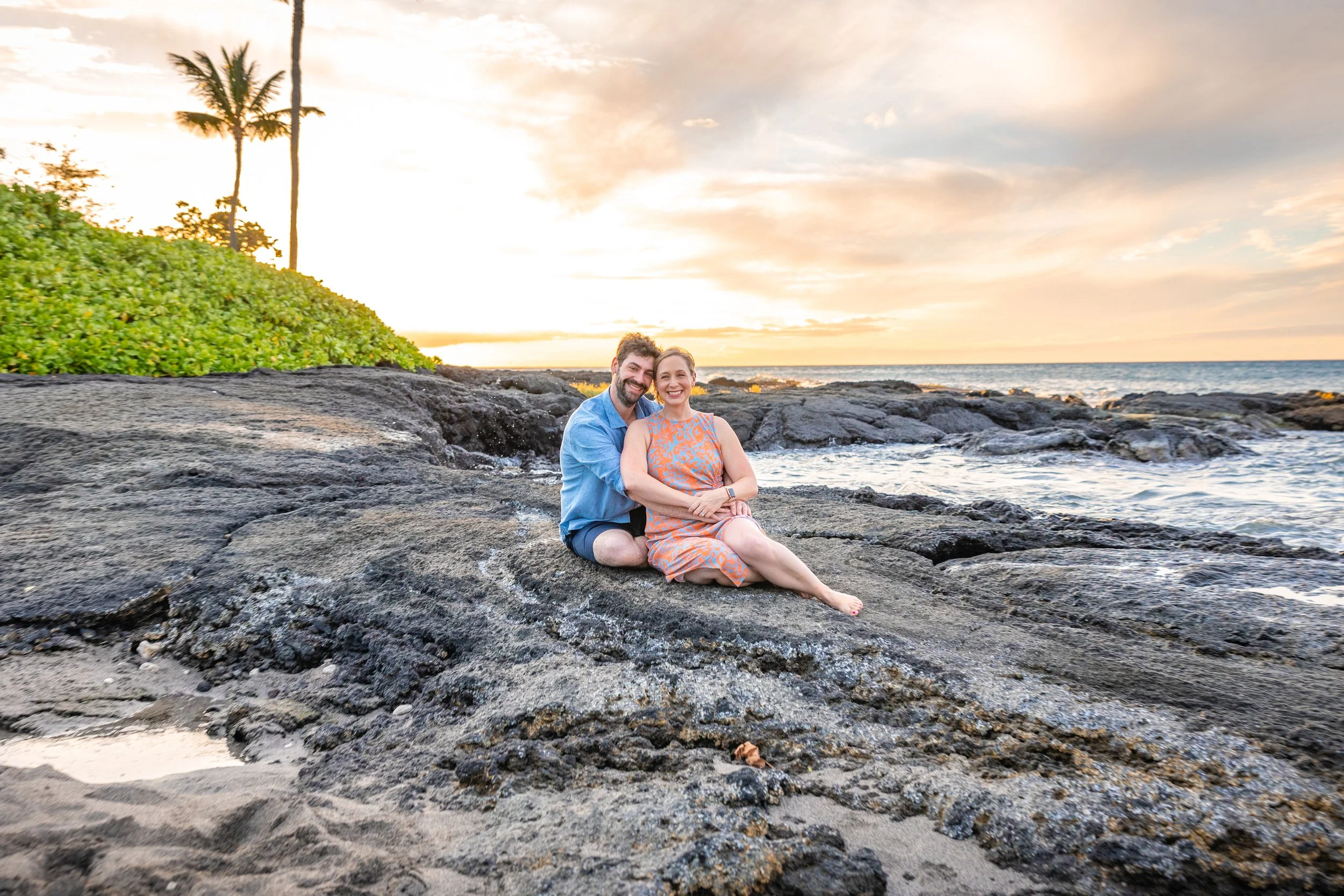 A smiling couple sitting on volcanic rocks by the beach during sunset, with palm trees and lush greenery in the background.