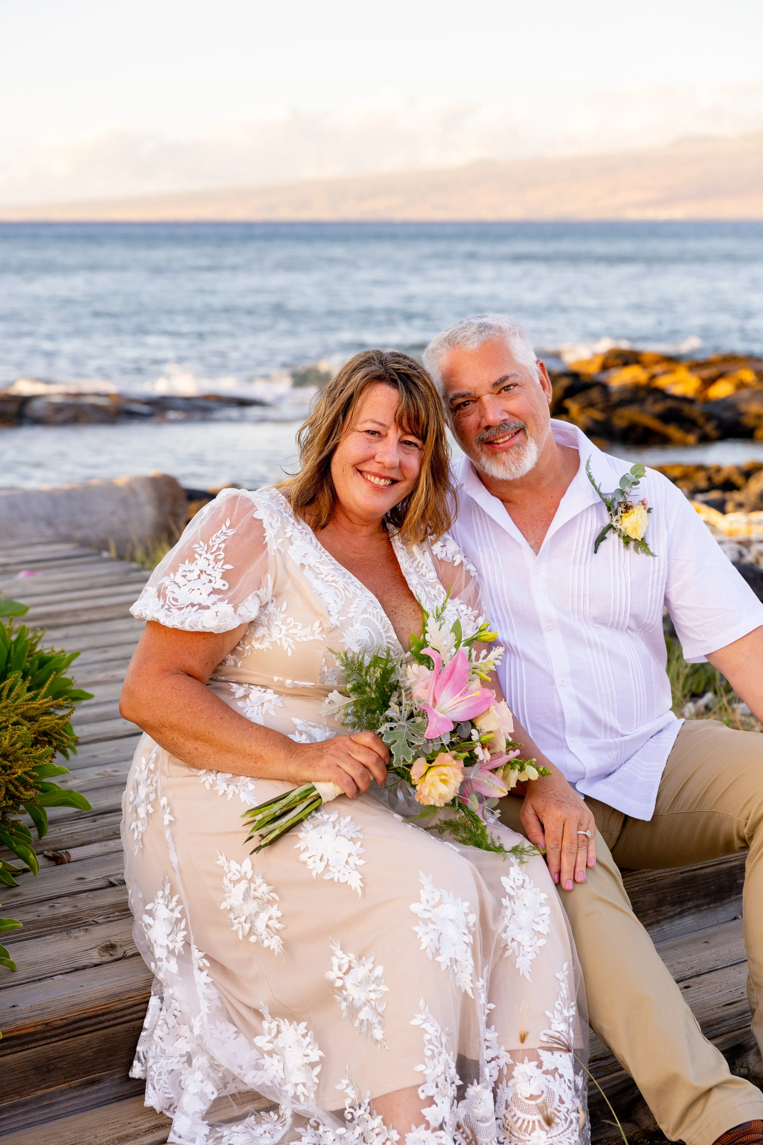 A smiling woman in a white lace wedding dress and a man in a white shirt and tan pants sitting together outdoors near the water, holding a bouquet of flowers.