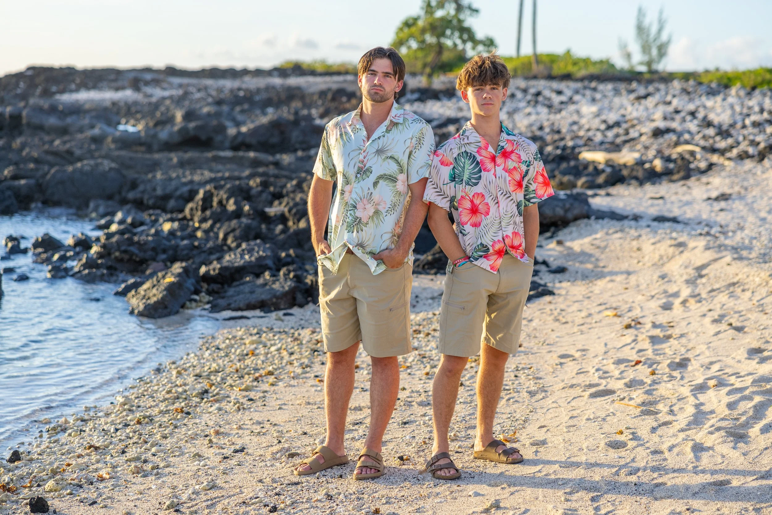 Two young men stand on a sandy beach with rocks and the ocean behind them, wearing floral Hawaiian shirts and shorts, looking at the camera.