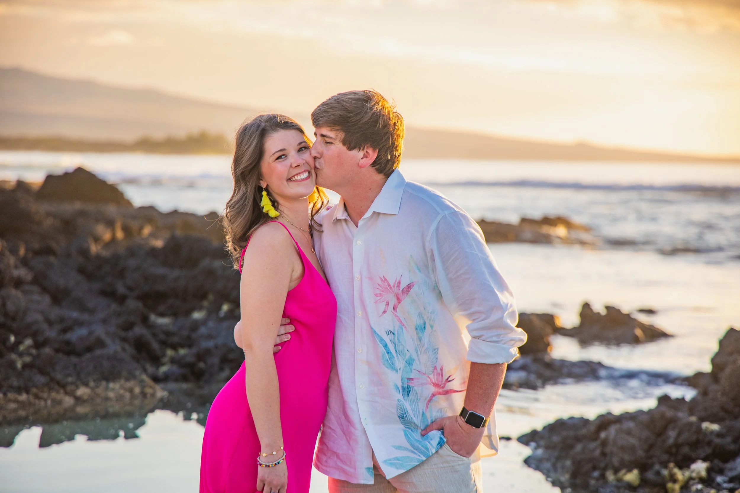 A young couple standing on rocky shoreline at sunset. The man is kissing the woman on the cheek while she smiles at the camera. The woman is wearing a bright pink dress and yellow earrings, and the man is in a white shirt with a tropical print. The o