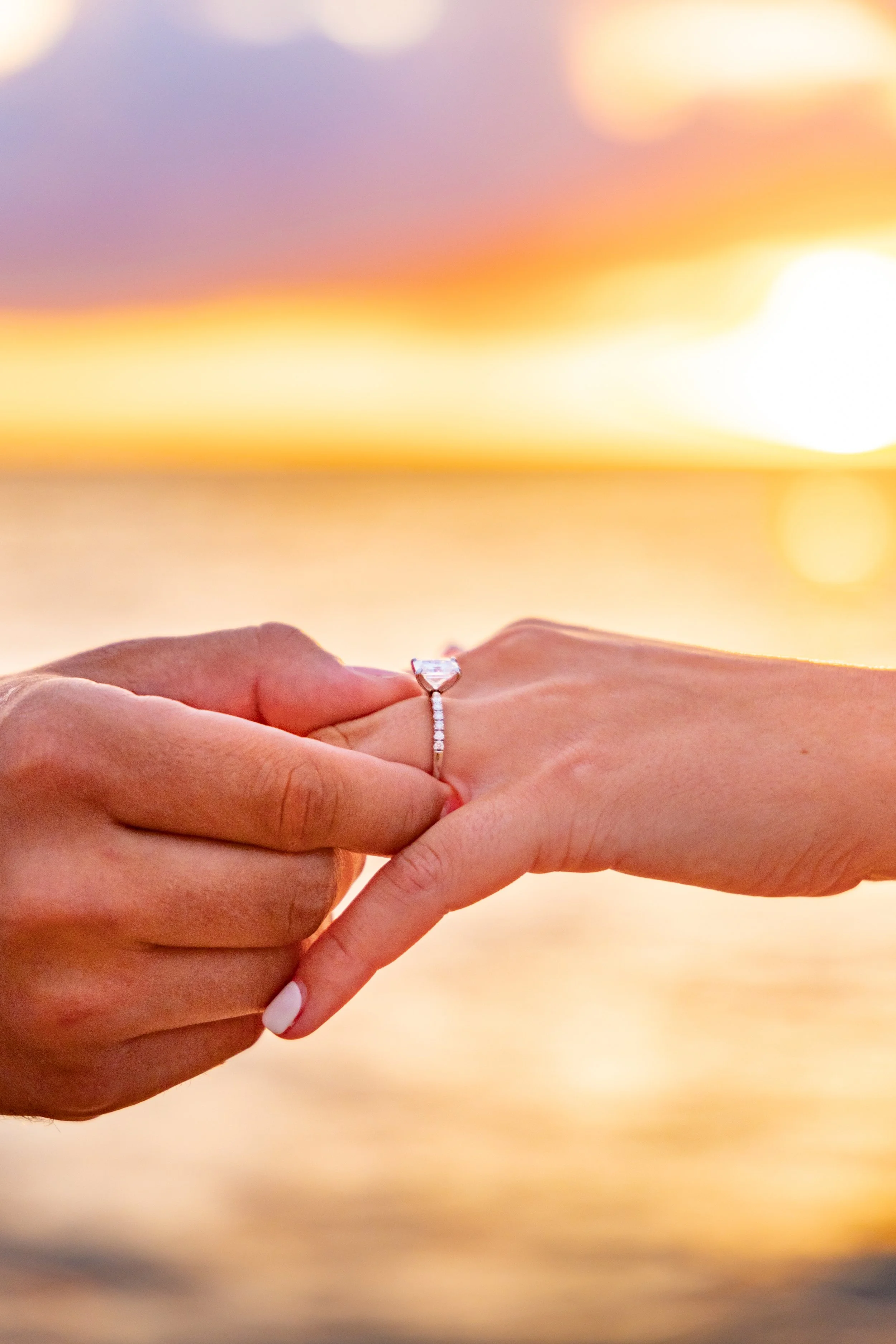 A person is placing an engagement ring on another person's finger during a sunset at the beach.