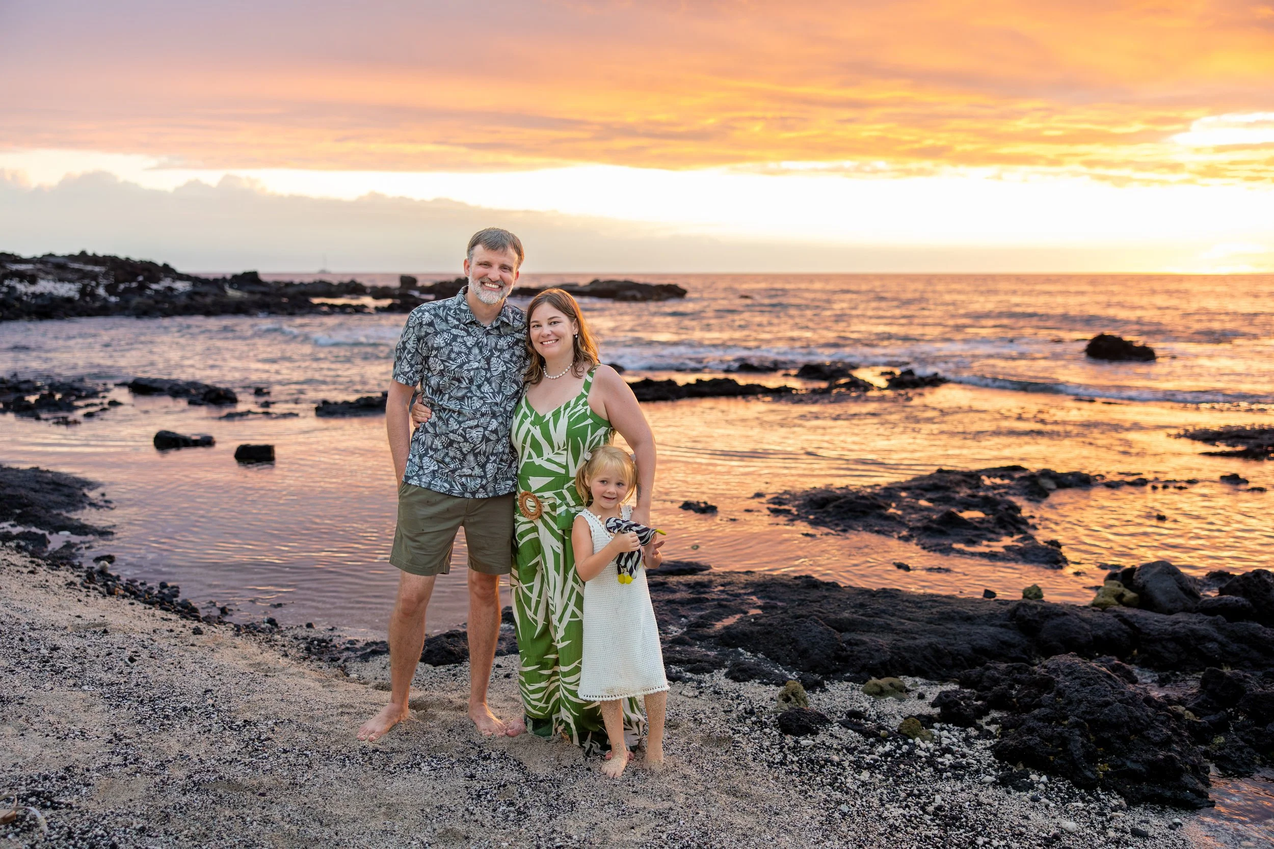 A family of three standing on a beach at sunset, with the ocean and sky in the background. The father, mother, and young girl are smiling.