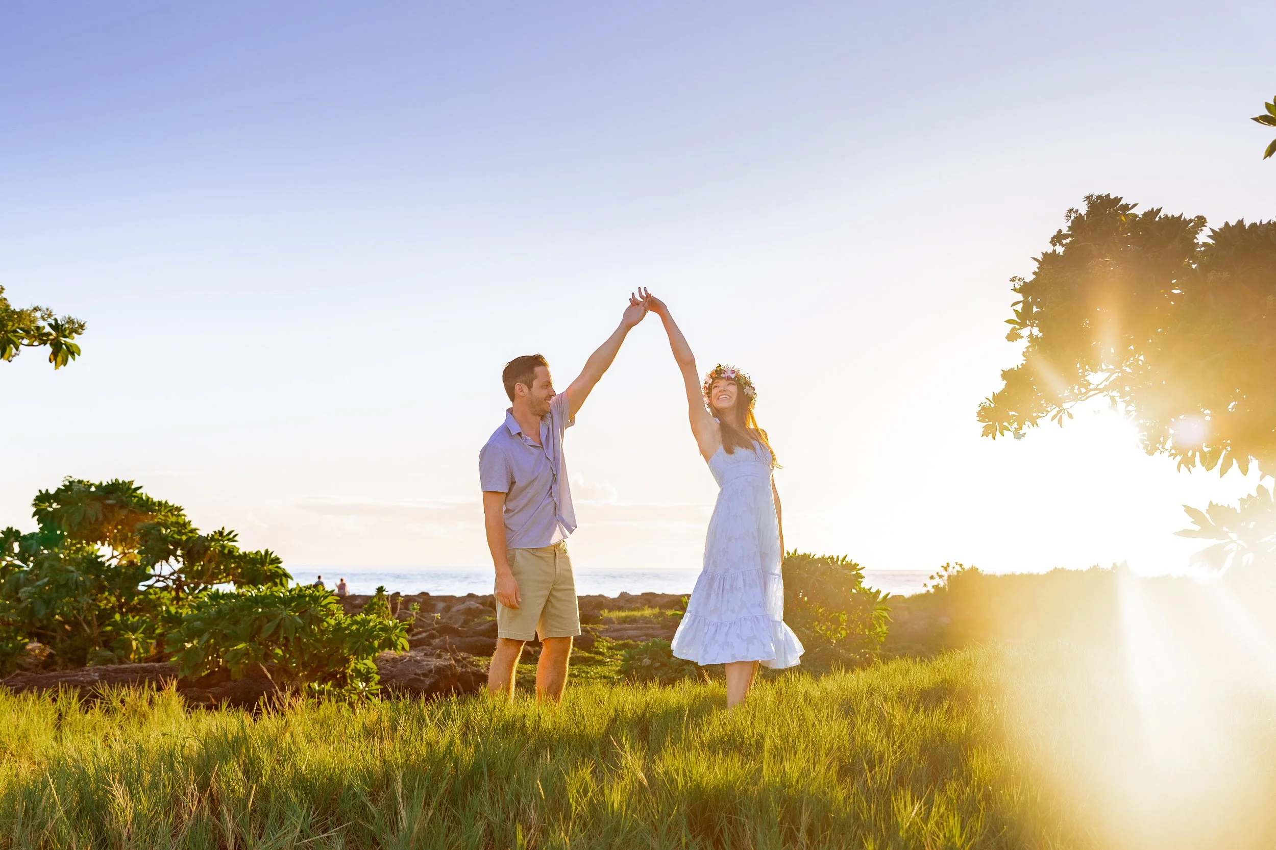 Couple enjoying a moment on a grassy field near a beach at sunset, with the man raising his hand to high-five the woman who is wearing a white dress and floral headband.
