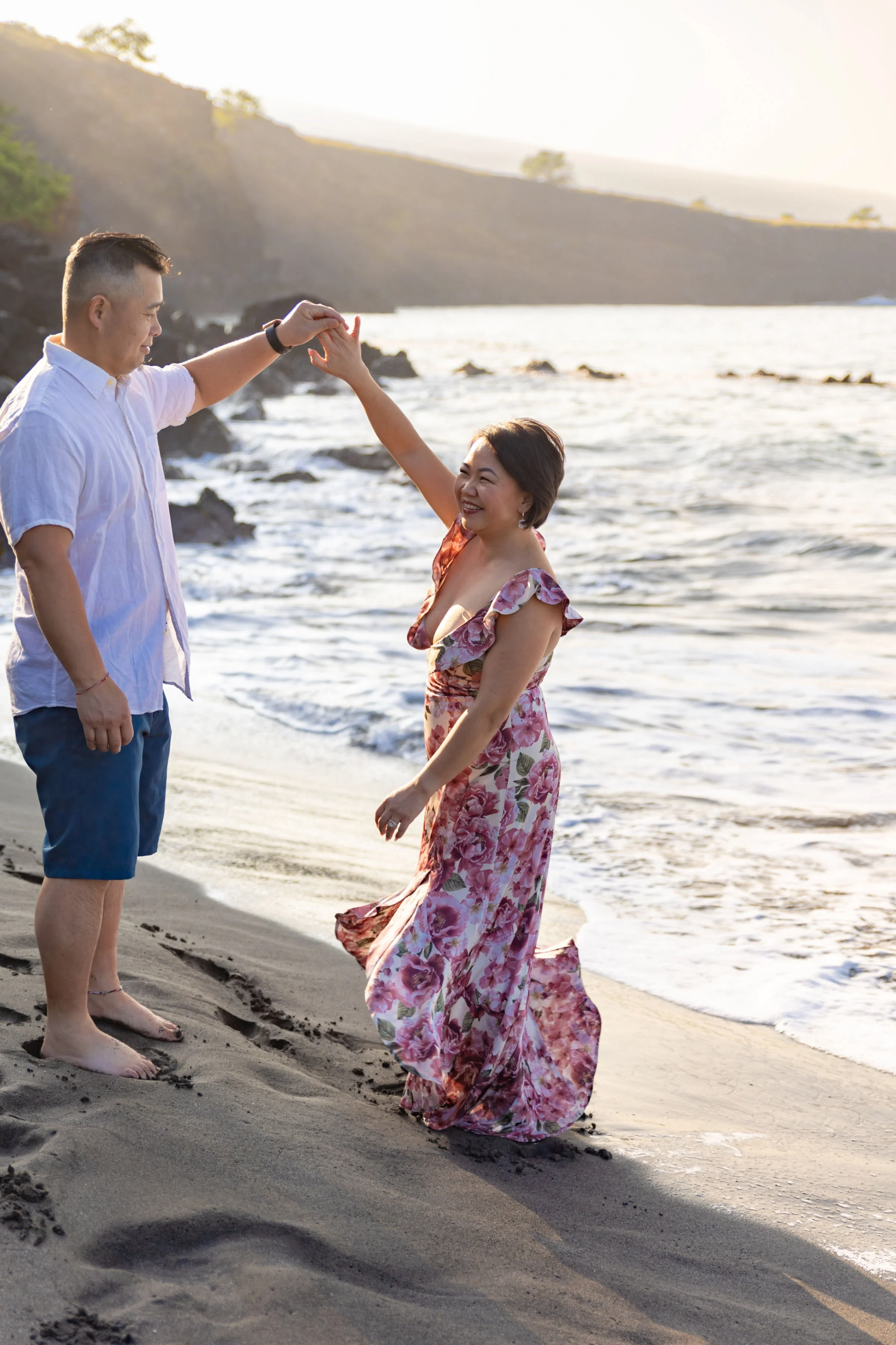A couple dancing on the beach during sunset, with the man holding the woman's hand as she twirls. The woman is wearing a floral dress, and the man is dressed in a white shirt and blue shorts. The ocean and rocky coastline are visible in the backgroun