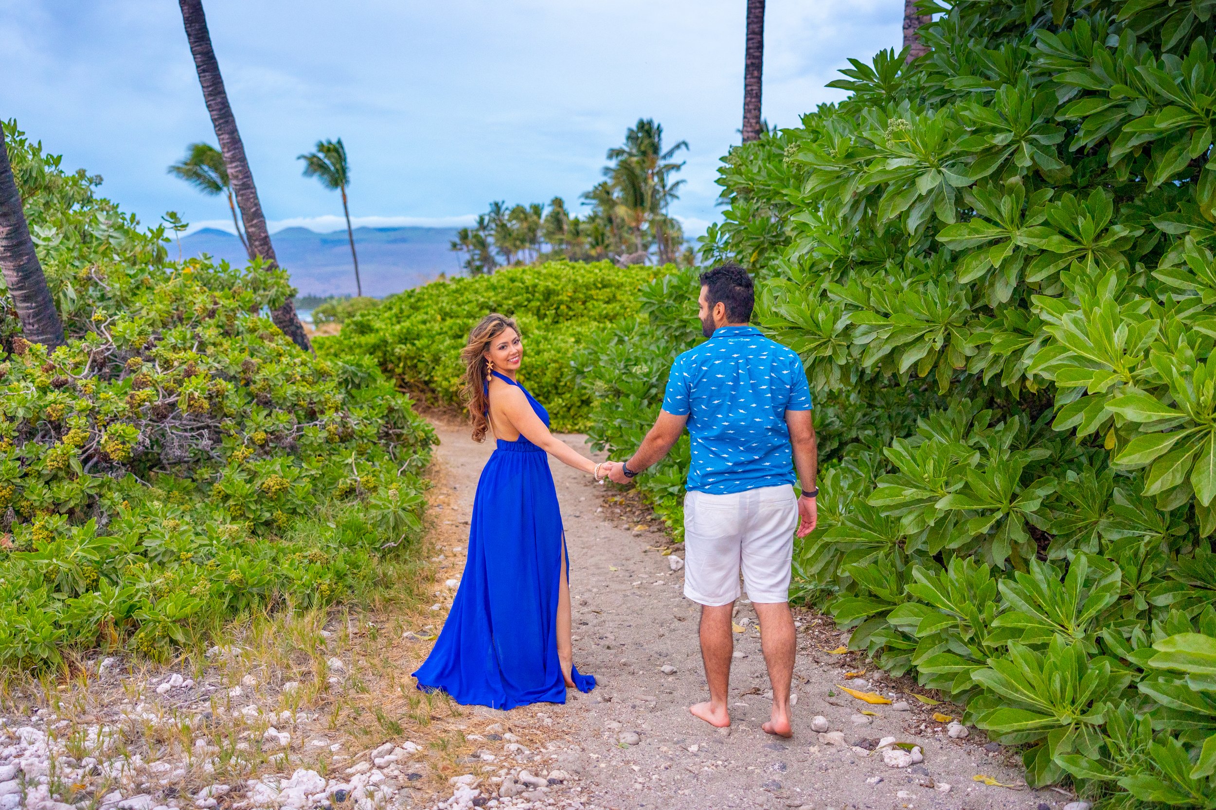 A couple holding hands on a dirt path through lush green foliage with palm trees in the background, near a beach or coastal area.