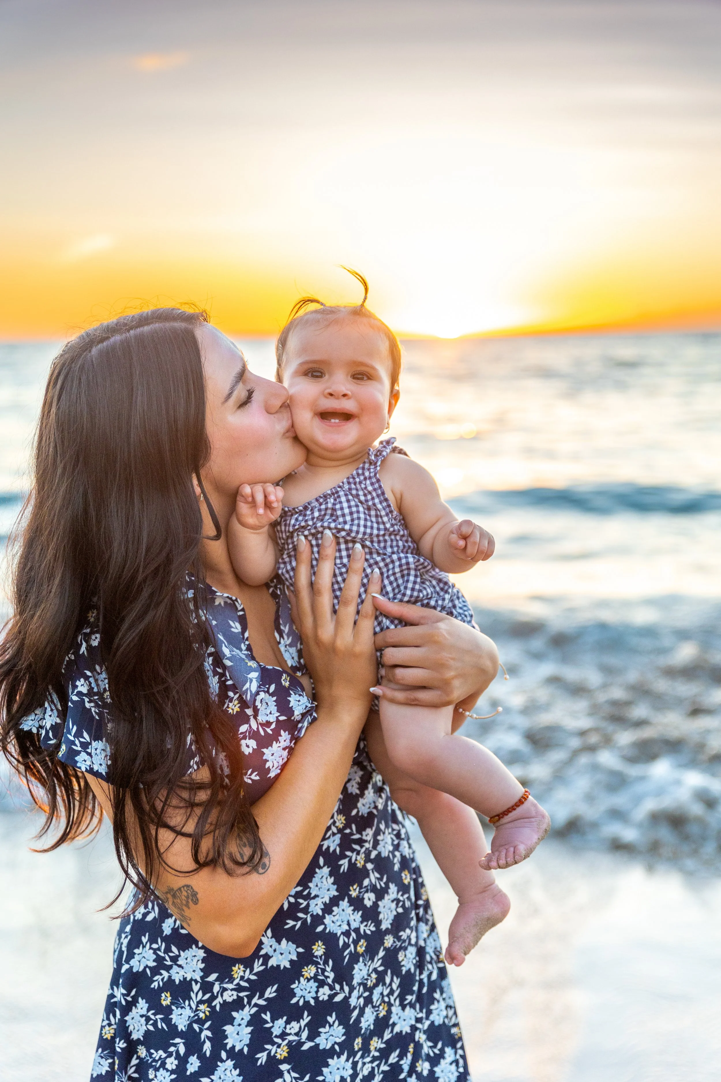A woman with long dark hair holding a smiling baby girl near the beach at sunset