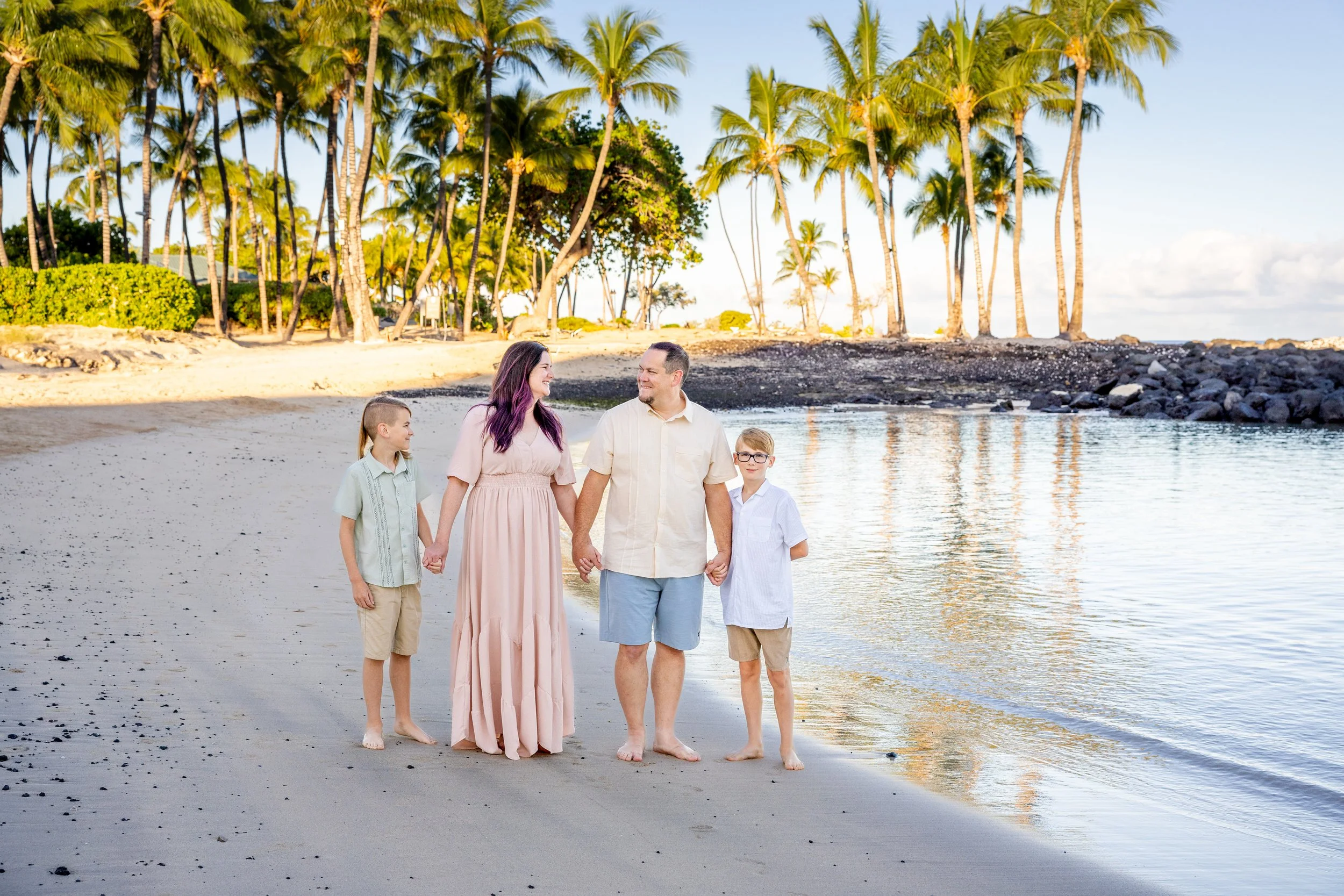 A family of four walking hand in hand along the beach, with palm trees in the background and calm water reflecting the sky.