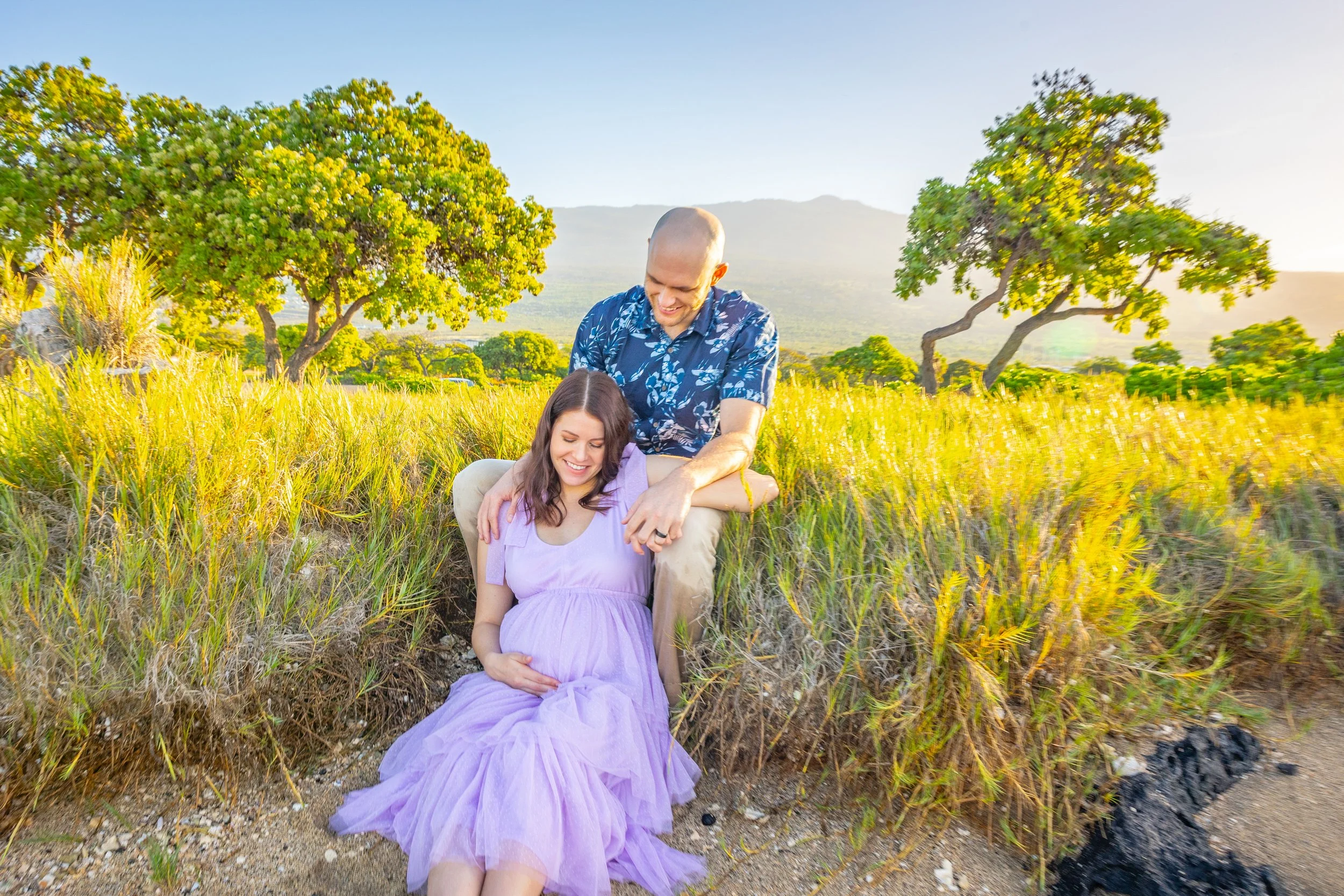 A smiling pregnant woman in a lavender dress sitting on the ground outdoors with a man standing behind her, placing his hand on her shoulder. They are in a grassy area with trees and mountains in the background during sunset.
