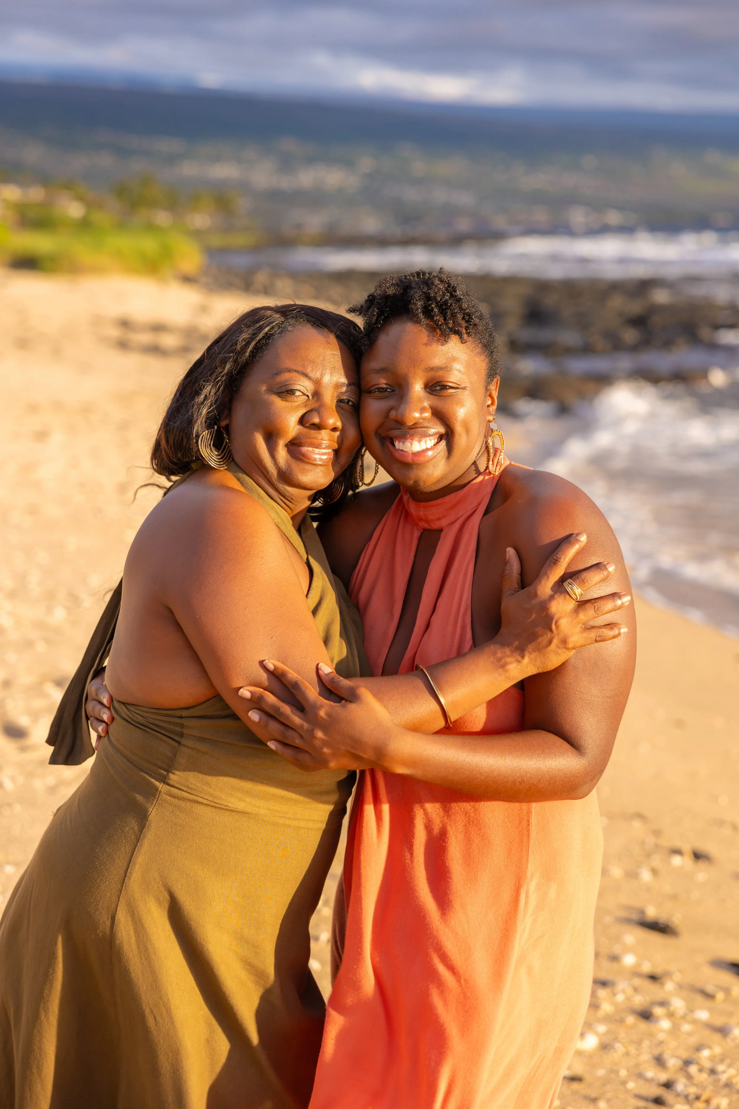 Two women hugging on a beach with waves and hills in the background, smiling warmly.
