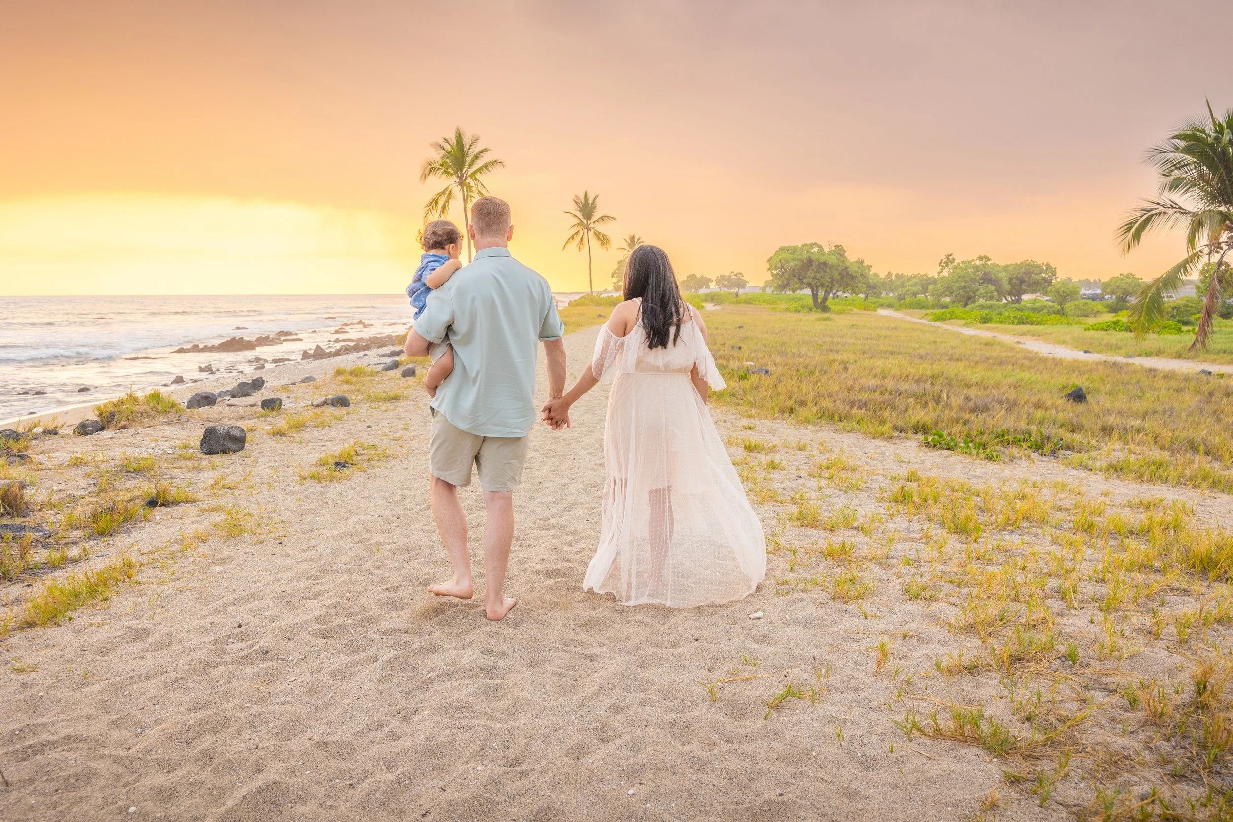 A family walking along a sandy beach at sunset, with the father holding a young child and the mother walking beside them, holding hands.
