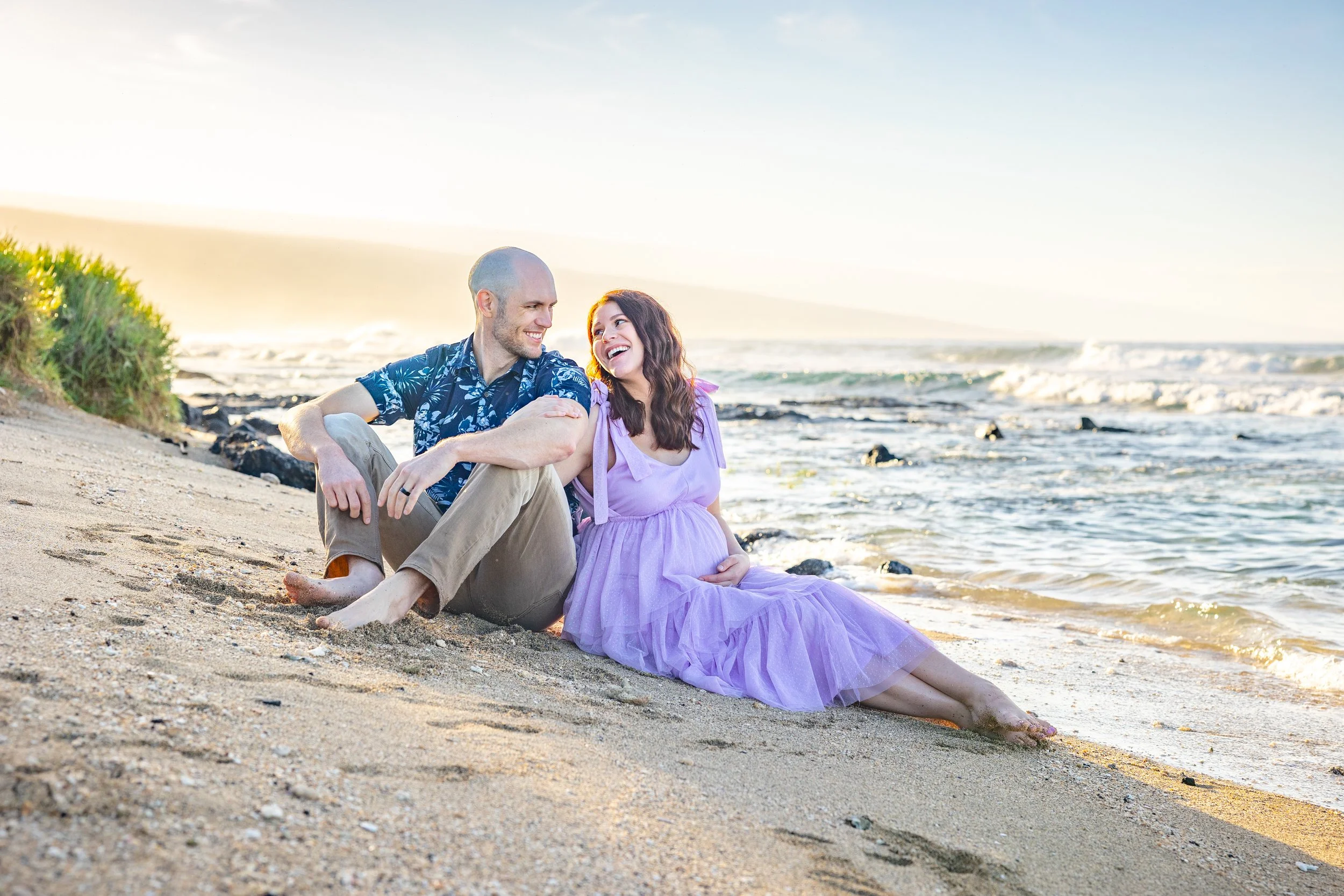 A smiling couple sitting on a sandy beach near the ocean, enjoying the sunset.
