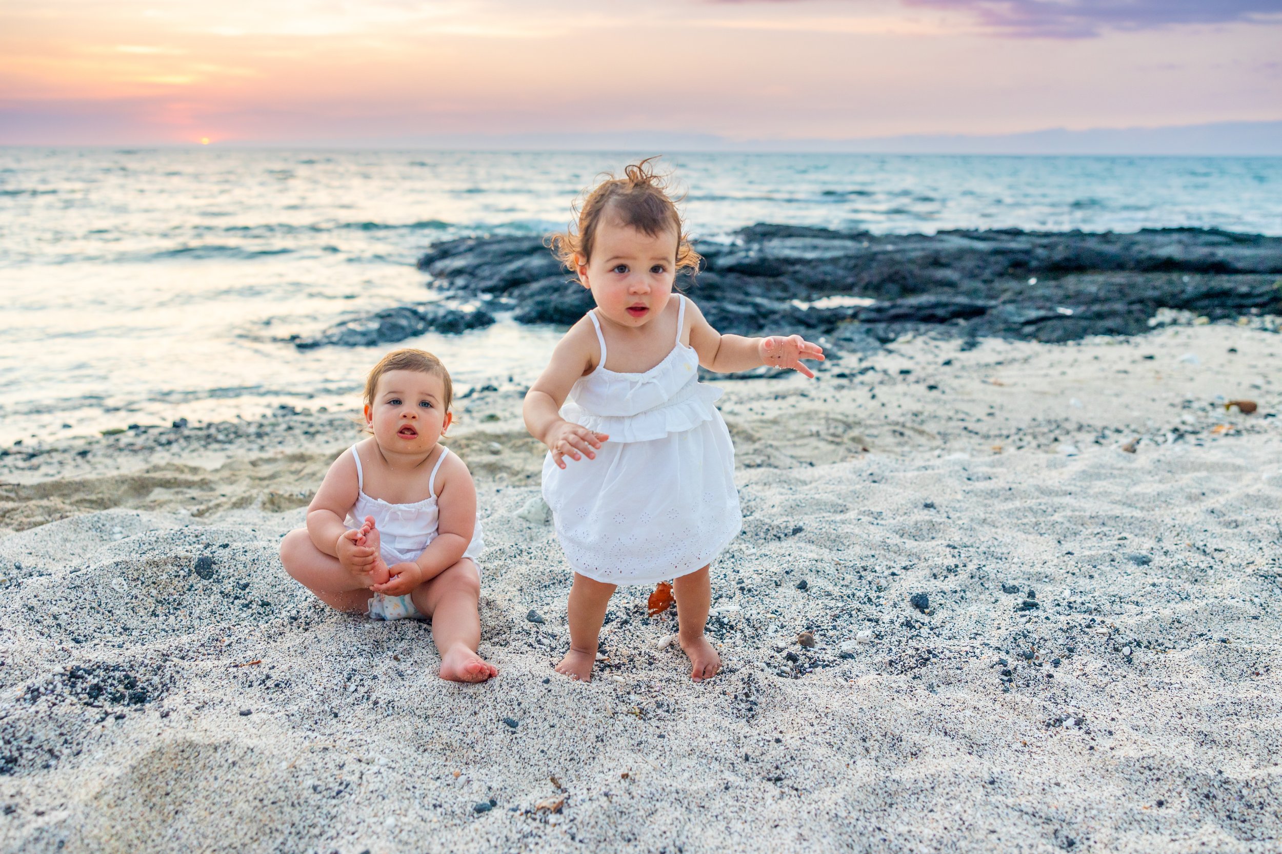 Two young children, one sitting and one standing, on a sandy beach at sunset with ocean waves and rocks in the background.