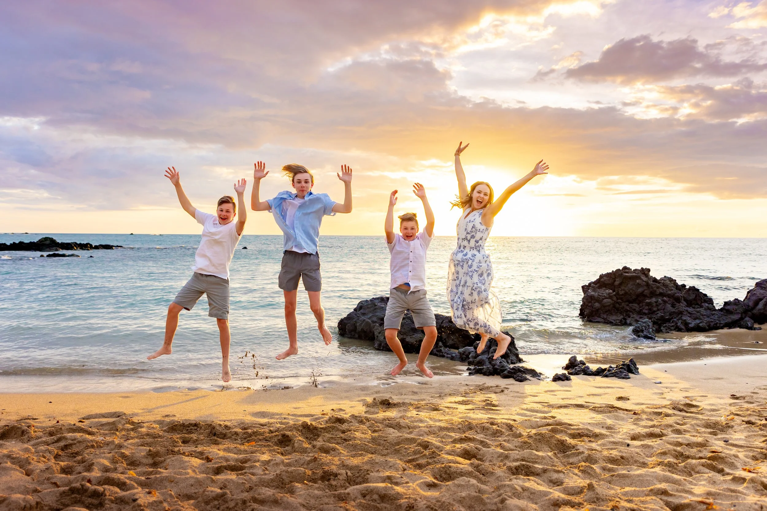 A happy family of four jumping on the beach at sunset, with rocks and ocean in the background.