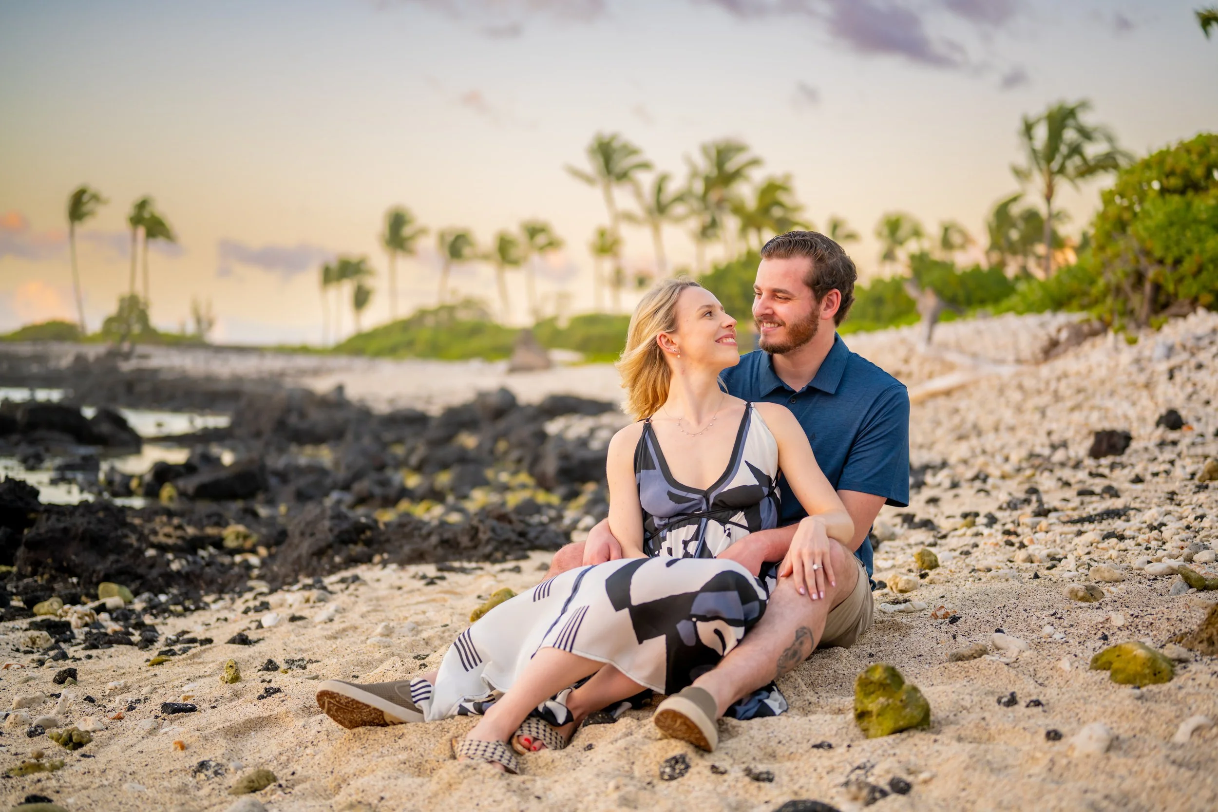 A couple sitting on a sandy beach at sunset, smiling and looking into each other's eyes, with palm trees and a cloudy sky in the background.