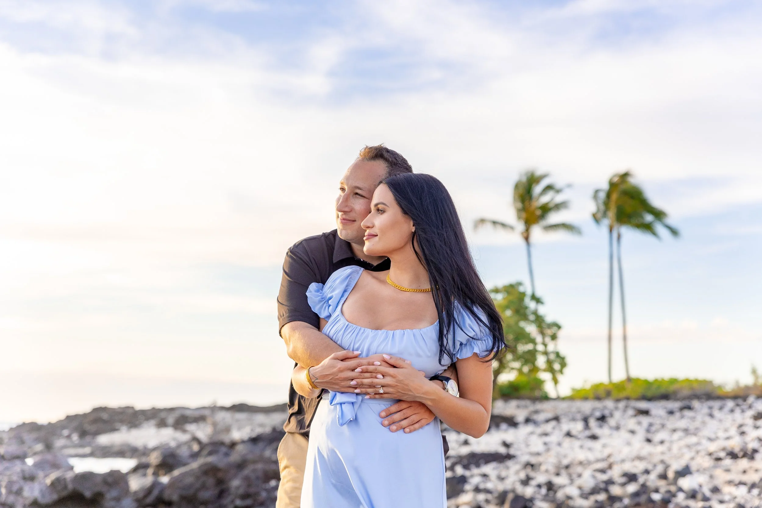 A couple standing on rocky ground at the beach with palm trees in the background, holding each other and gazing into the distance during a sunset or sunrise.