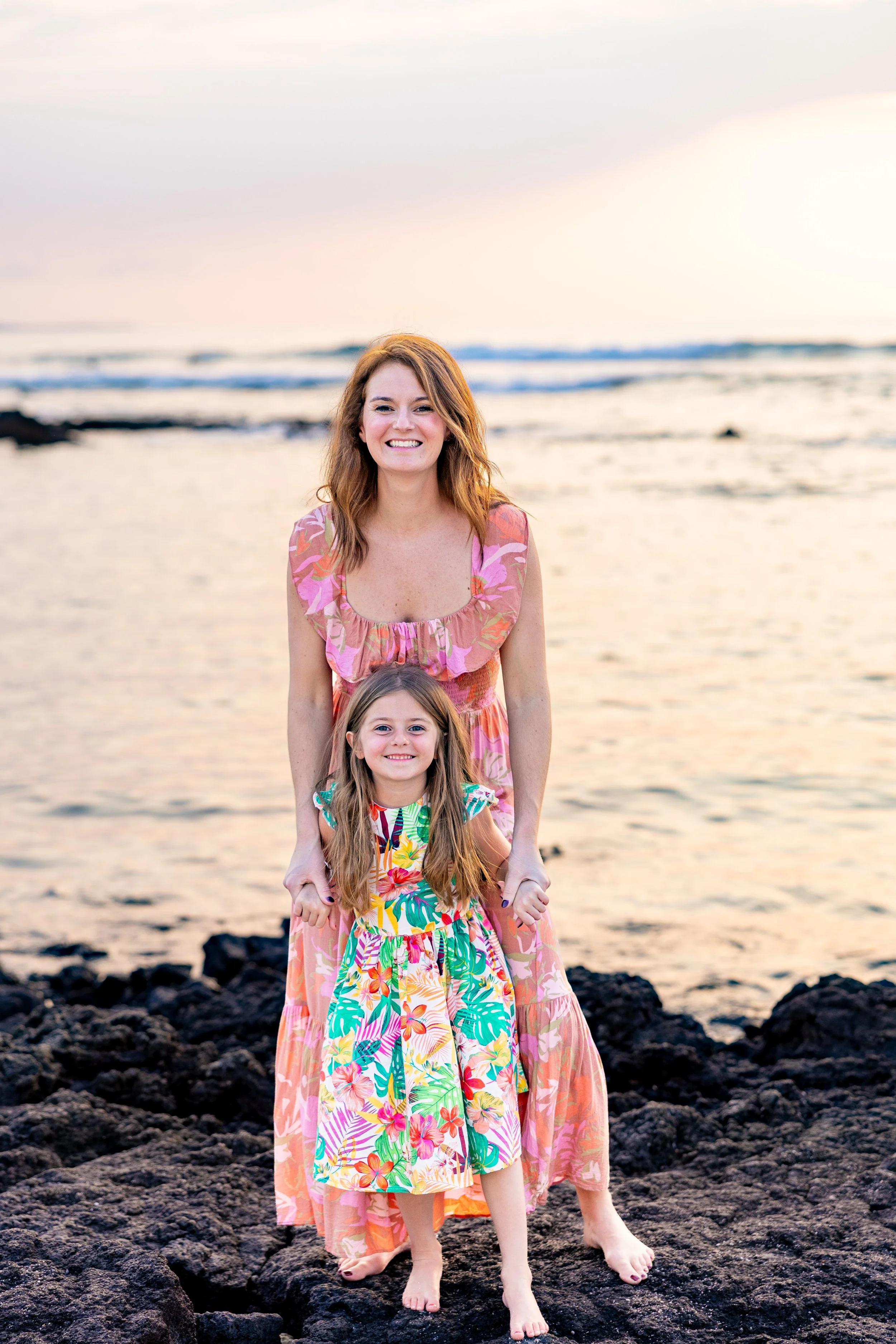 A woman and a young girl stand barefoot on rocky terrain at the beach during sunset, holding hands and smiling at the camera.