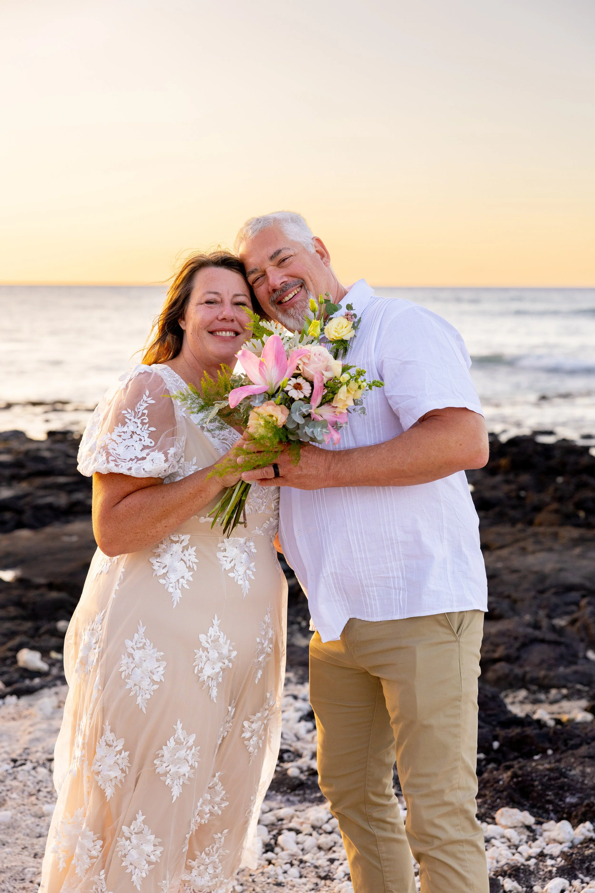 A happy couple on the beach at sunset, with the woman holding a bouquet of pink and white flowers, both smiling and embracing.