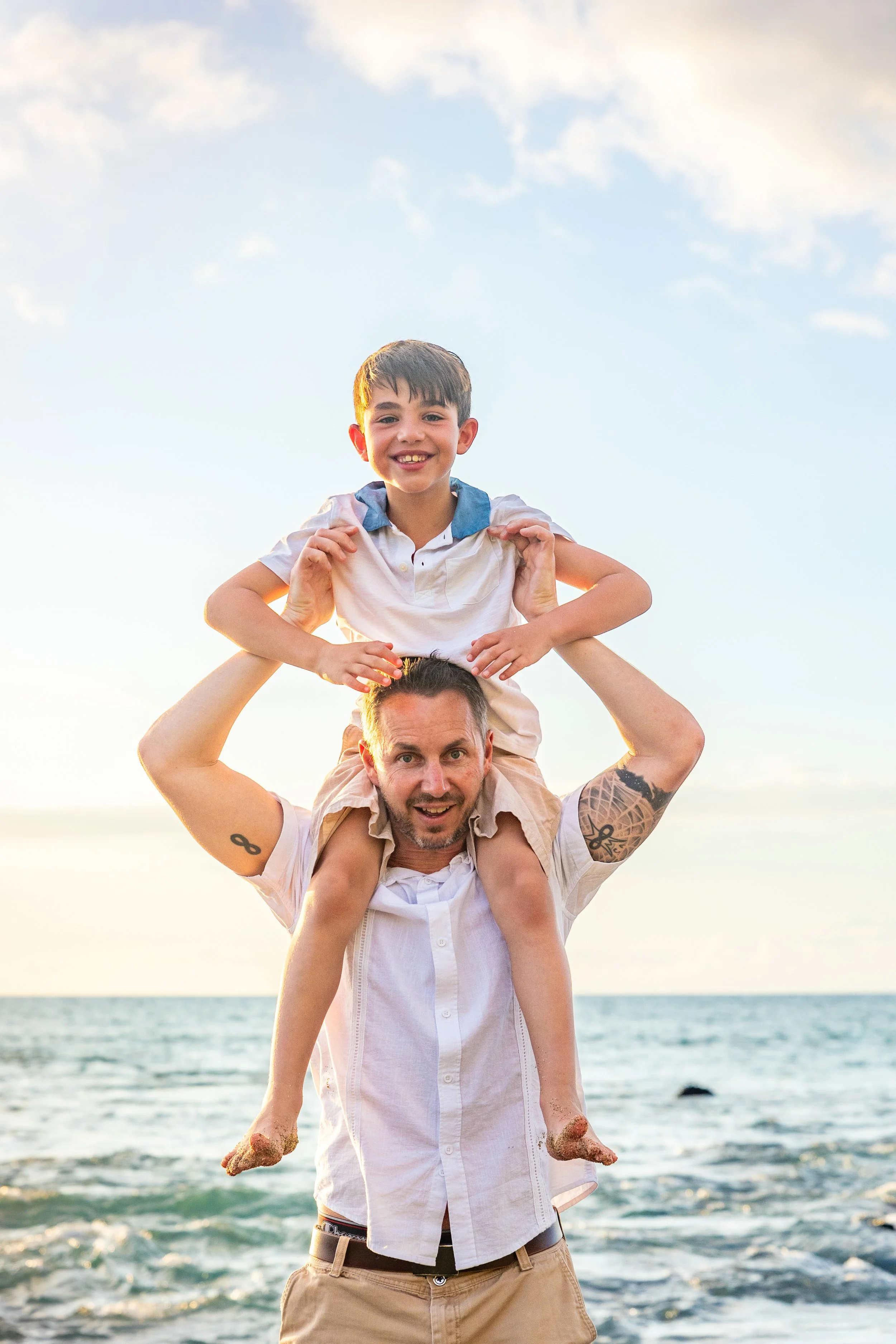 A man with tattoos on his arms carrying a young boy on his shoulders at the beach during sunset, both smiling.