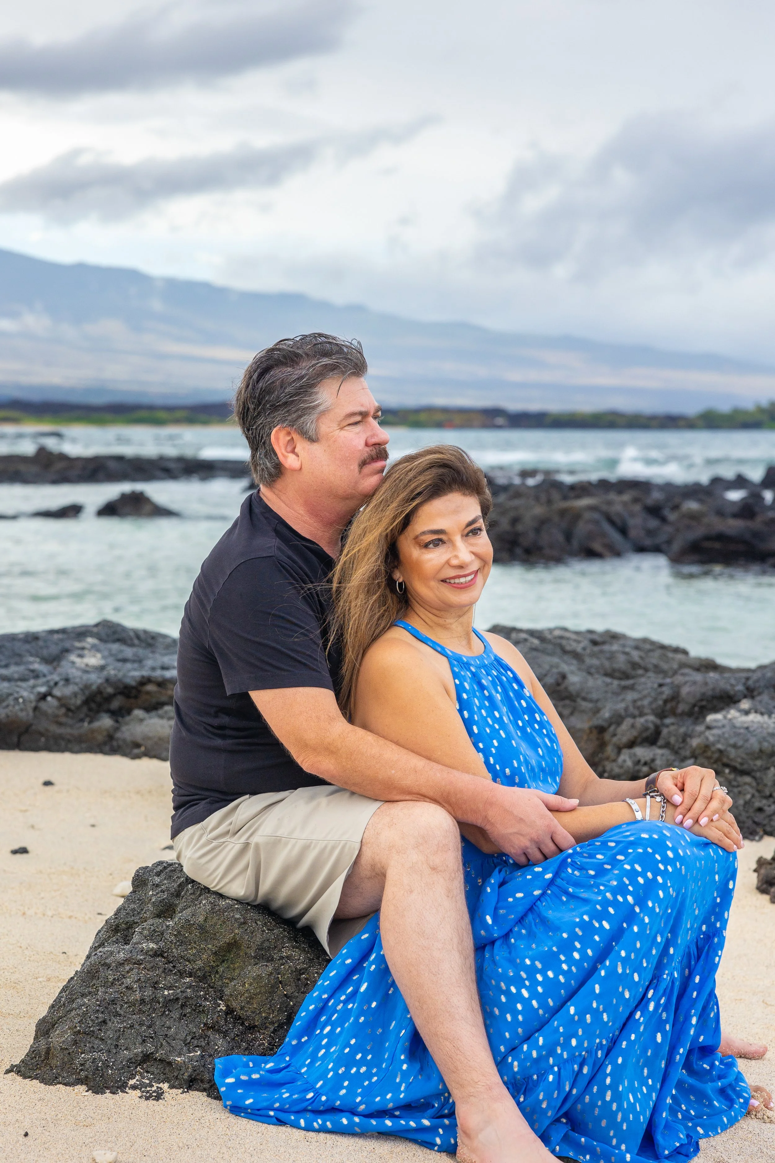 A couple sitting on a rock at the beach with the ocean and mountains in the background.