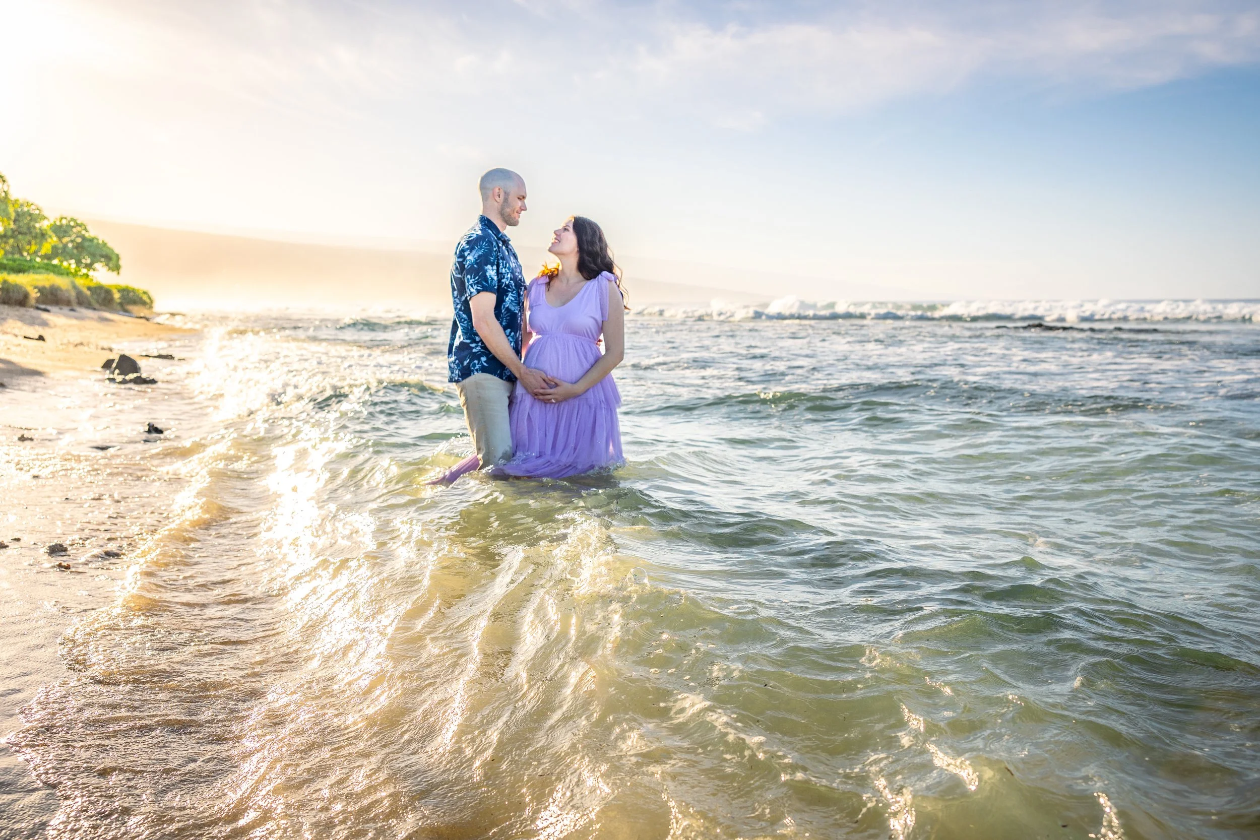 A couple standing in the ocean with waves, holding hands, looking at each other during sunset or sunrise, on a beach with some greenery in the background.