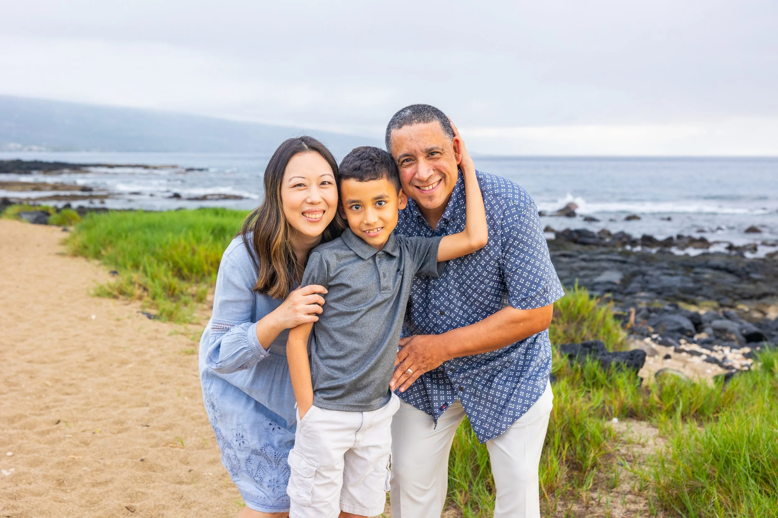 A family of three smiling and hugging on a beach with ocean and cloudy sky in the background