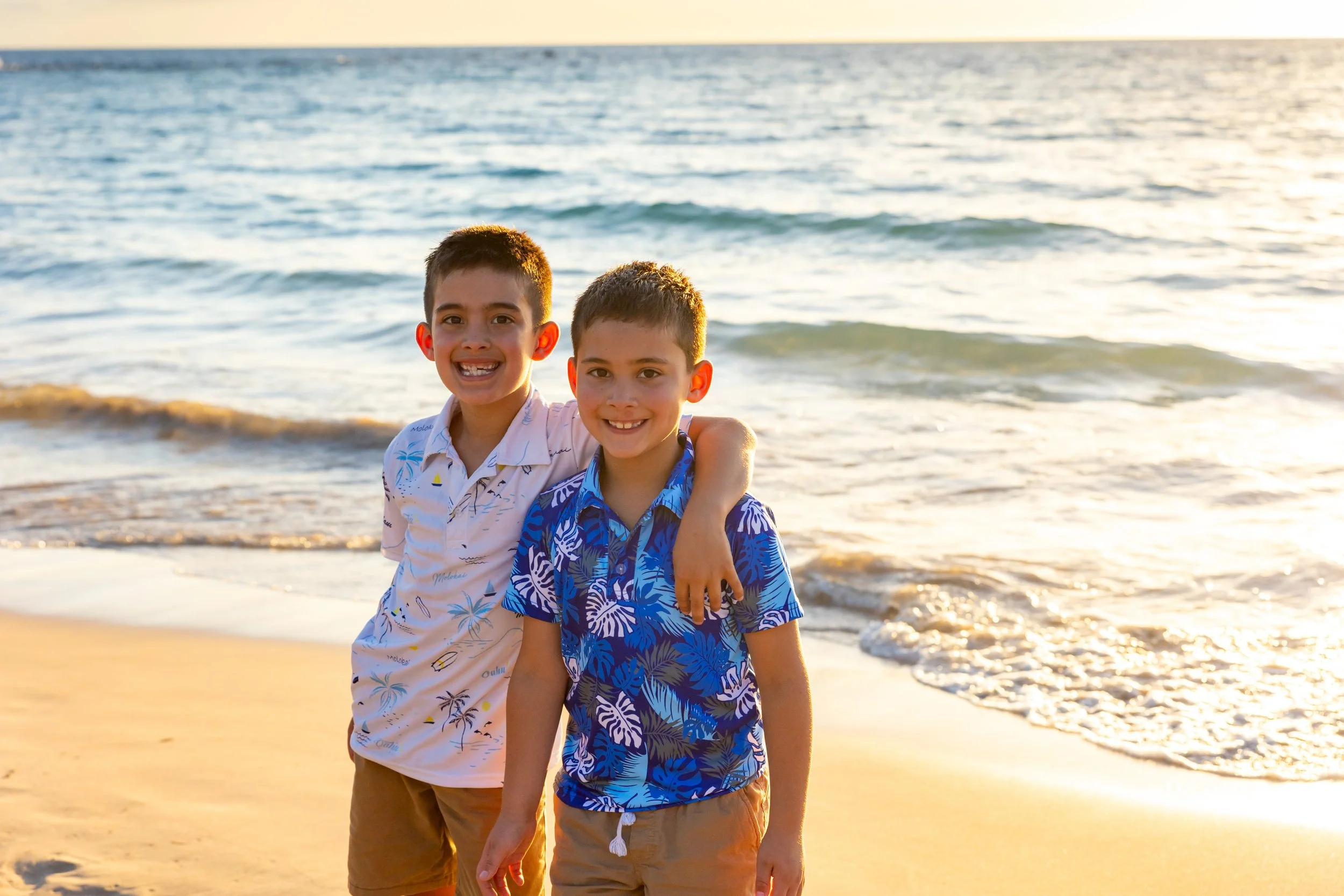 Two young boys smiling at the beach during sunset with ocean waves in the background.