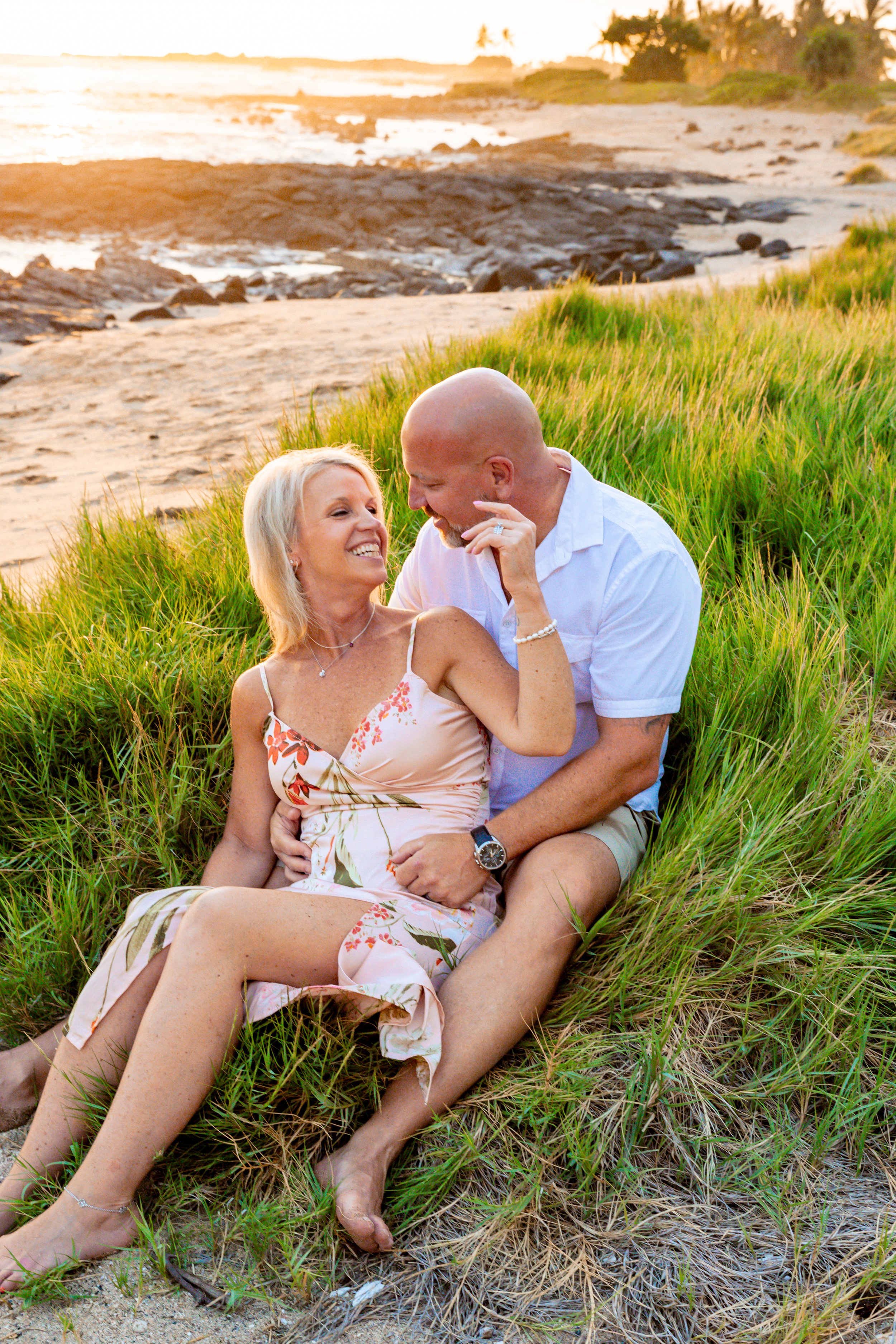 A couple sitting on the grass by the beach during sunset, smiling and looking at each other.